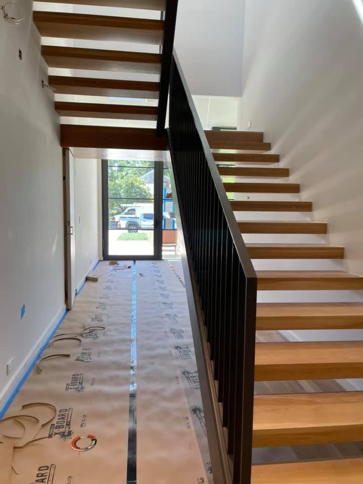Interior view of a staircase with wooden steps and black metal railing, with a glass door at the end showing a parked truck and trees outside, and a floor covered in protective paper.