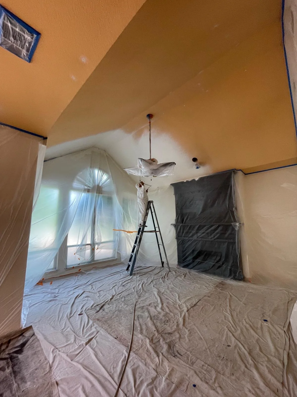 A room under renovation with plastic sheets covering the walls and floor, a stepladder, a person painting or working on the ceiling, and a window with curtains.