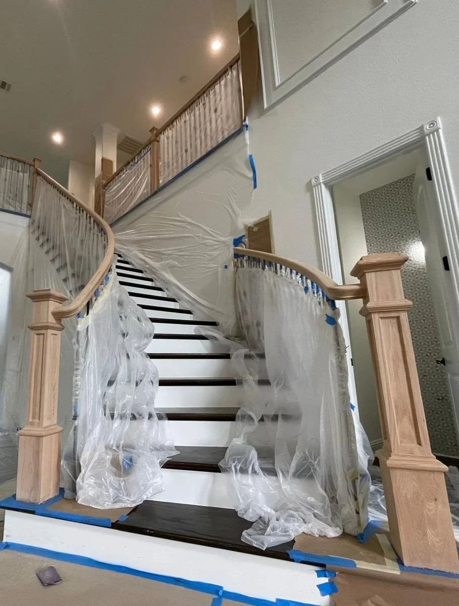 A staircase under renovation with plastic sheets and painter's tape. The wooden handrails and new dark wood treads are visible, and the area is protected for painting or refinishing.