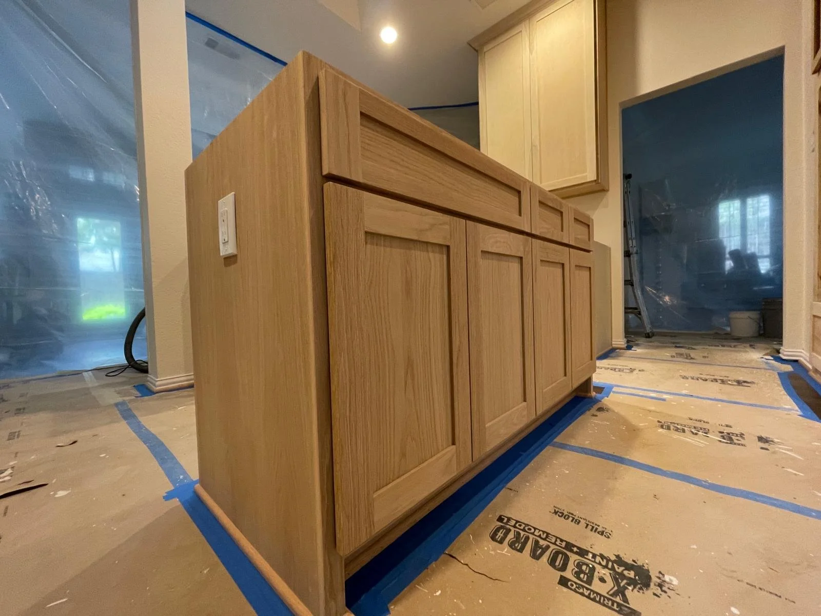 A kitchen island with wooden cabinets in a room under construction, with protective plastic coverings and blue painter's tape on the floor.