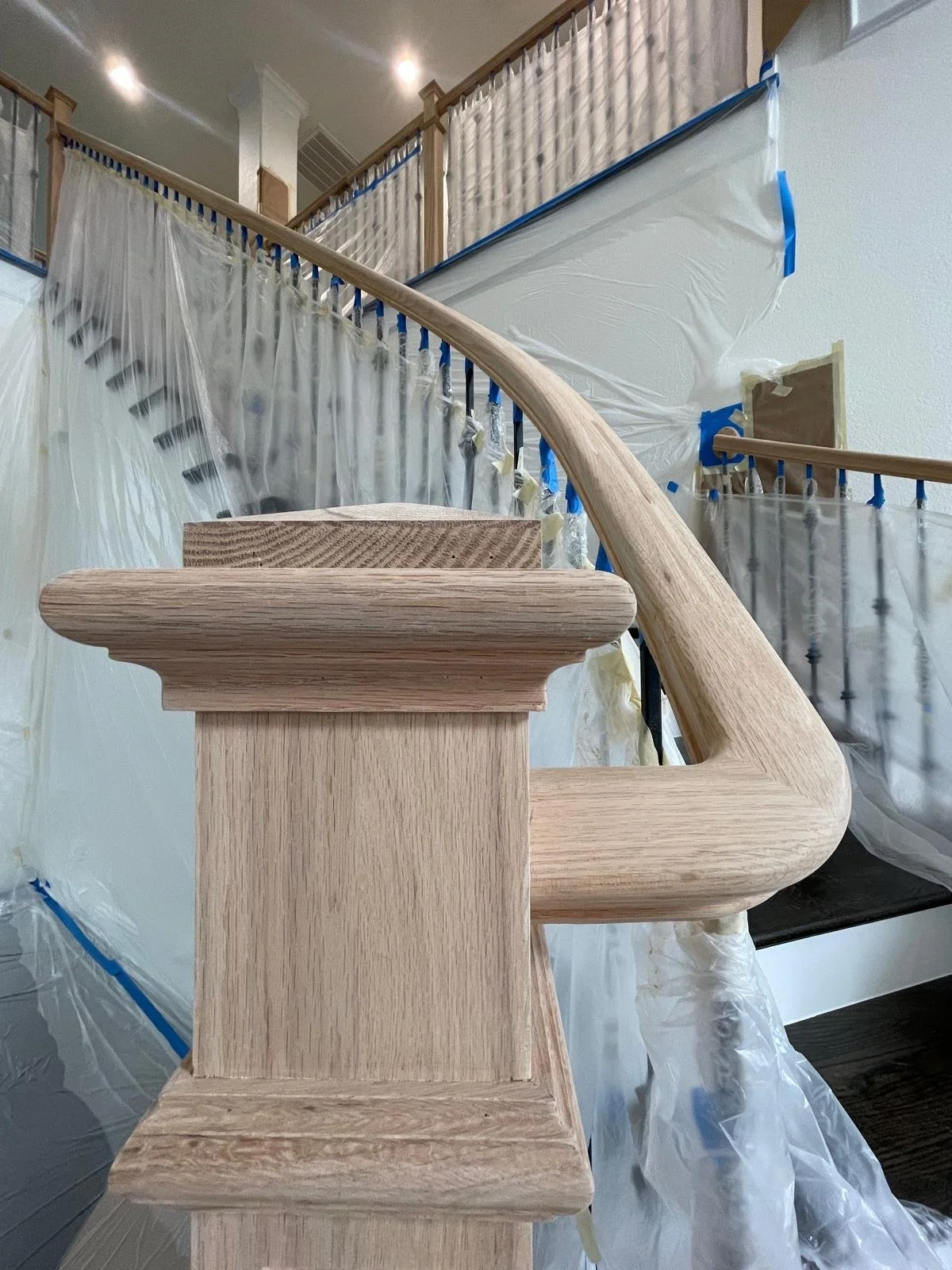 Close-up of a new wooden handrail and newel post on a staircase under construction, with plastic sheeting and painter's tape in the background.