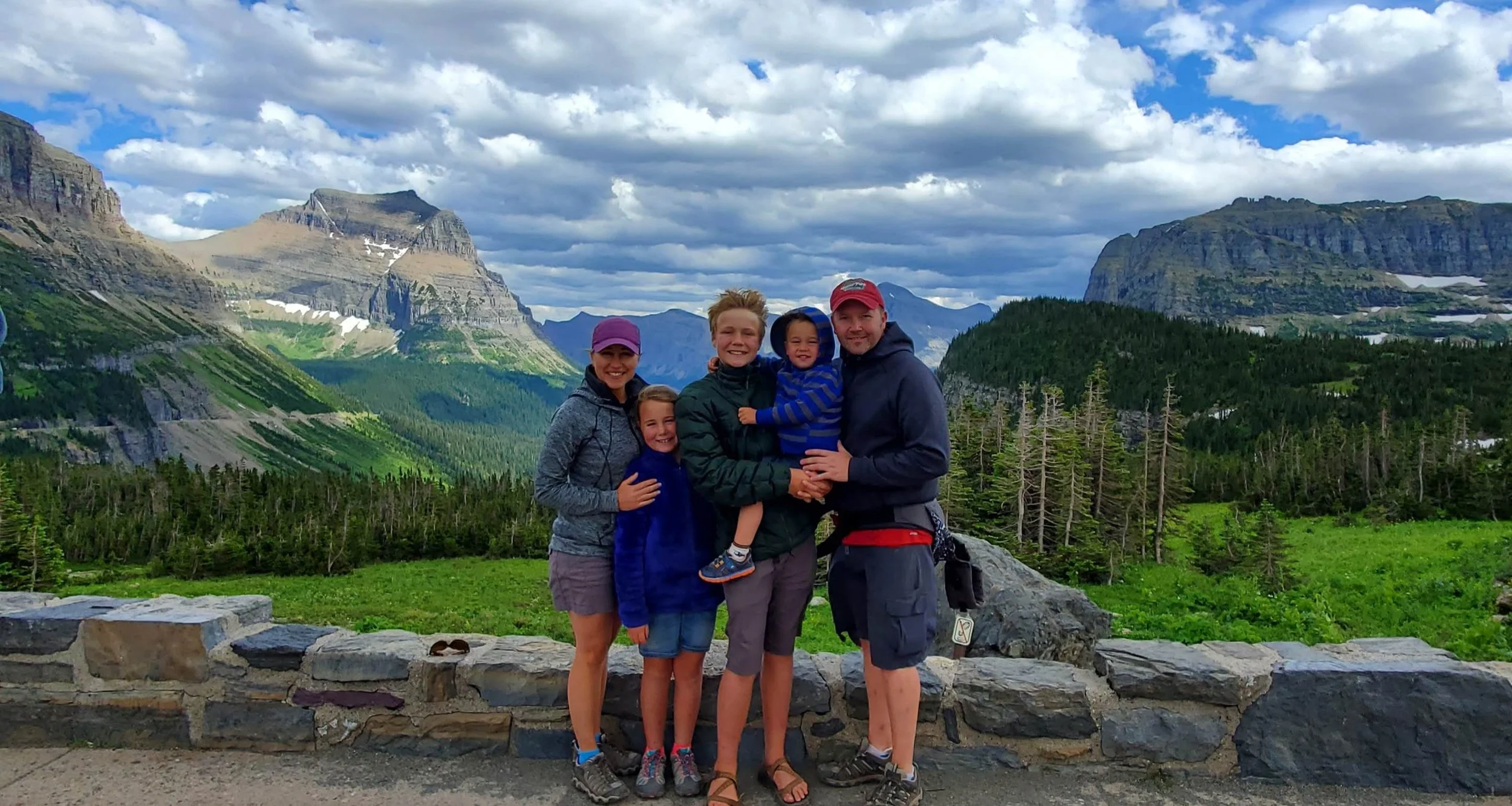 Dianne Morgan, owner of MorTravel and her Family of five posing together outdoors against a mountainous landscape with green forests, rocky peaks, and partly cloudy sky.