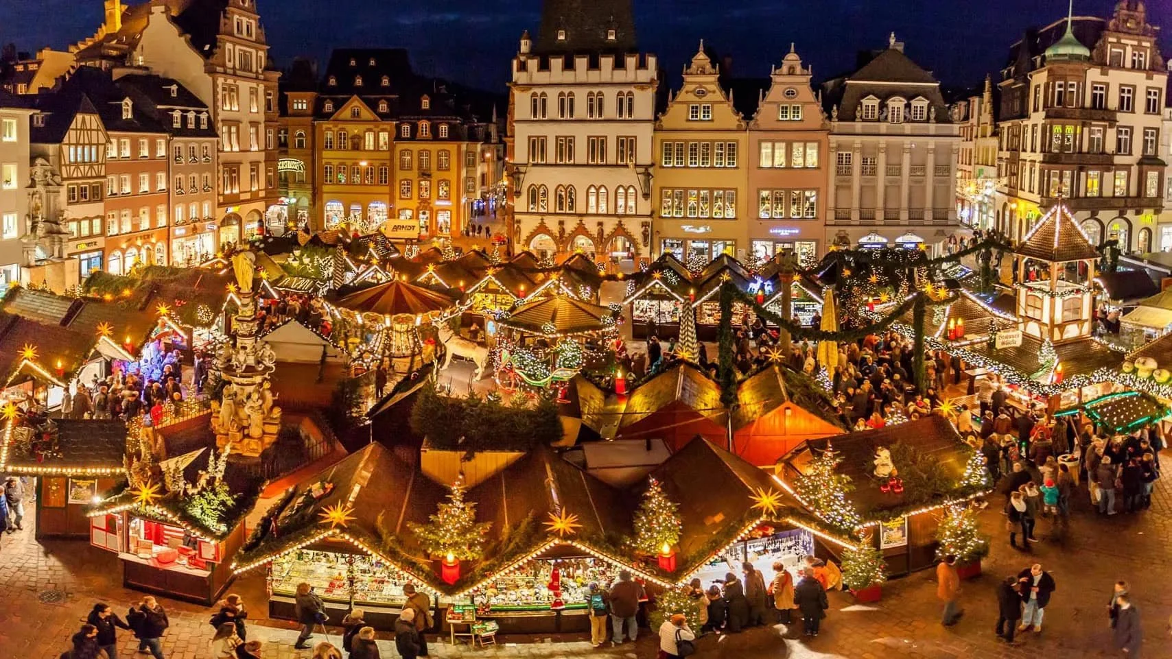 Nighttime scene of a Christmas market in a European town square with decorated stalls, Christmas trees, and crowds of people.  River cruises are very popular in Europe and stop at these christmas markets.