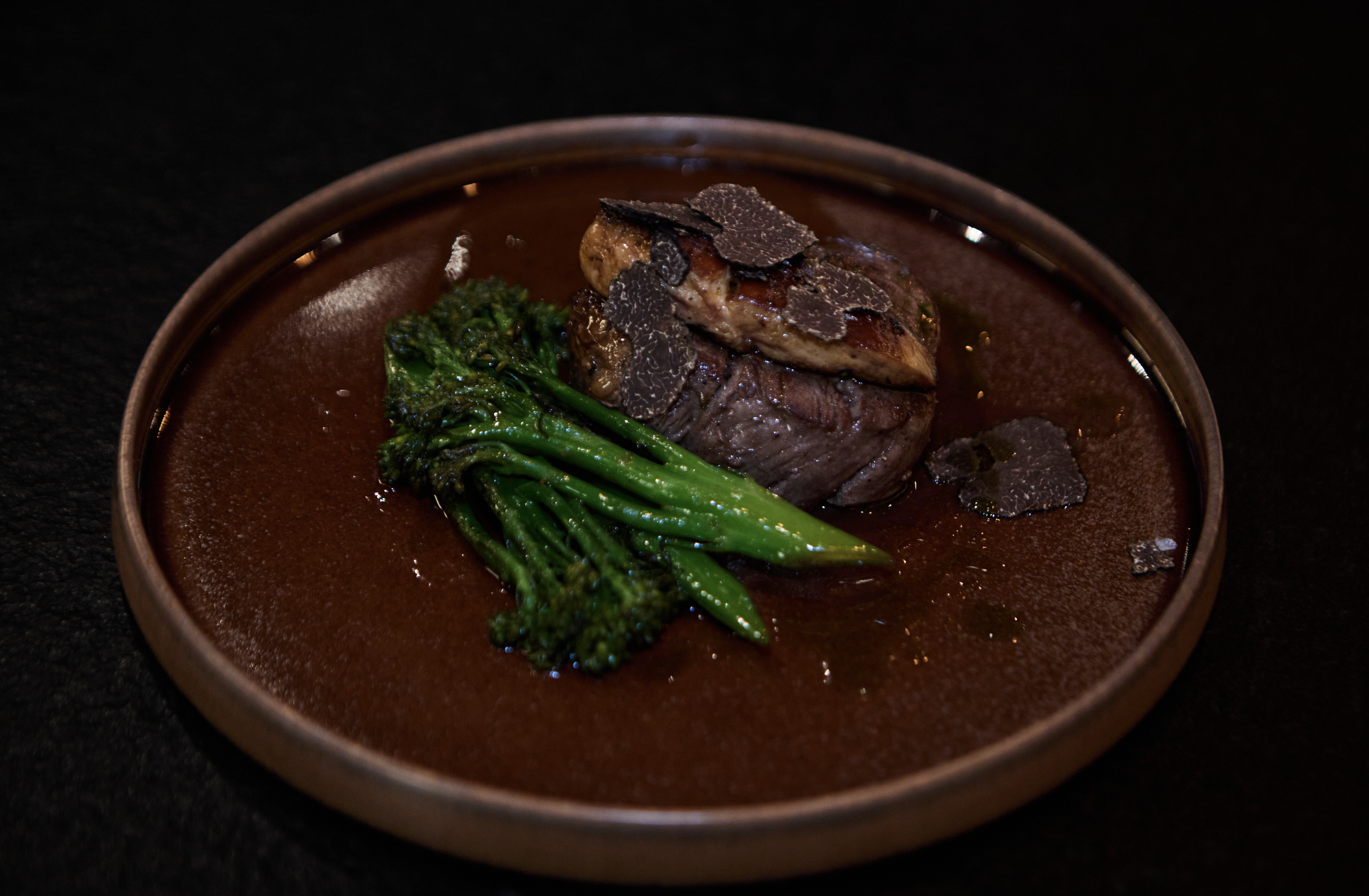 A plated gourmet steak with black truffles and broccolini on a dark surface.