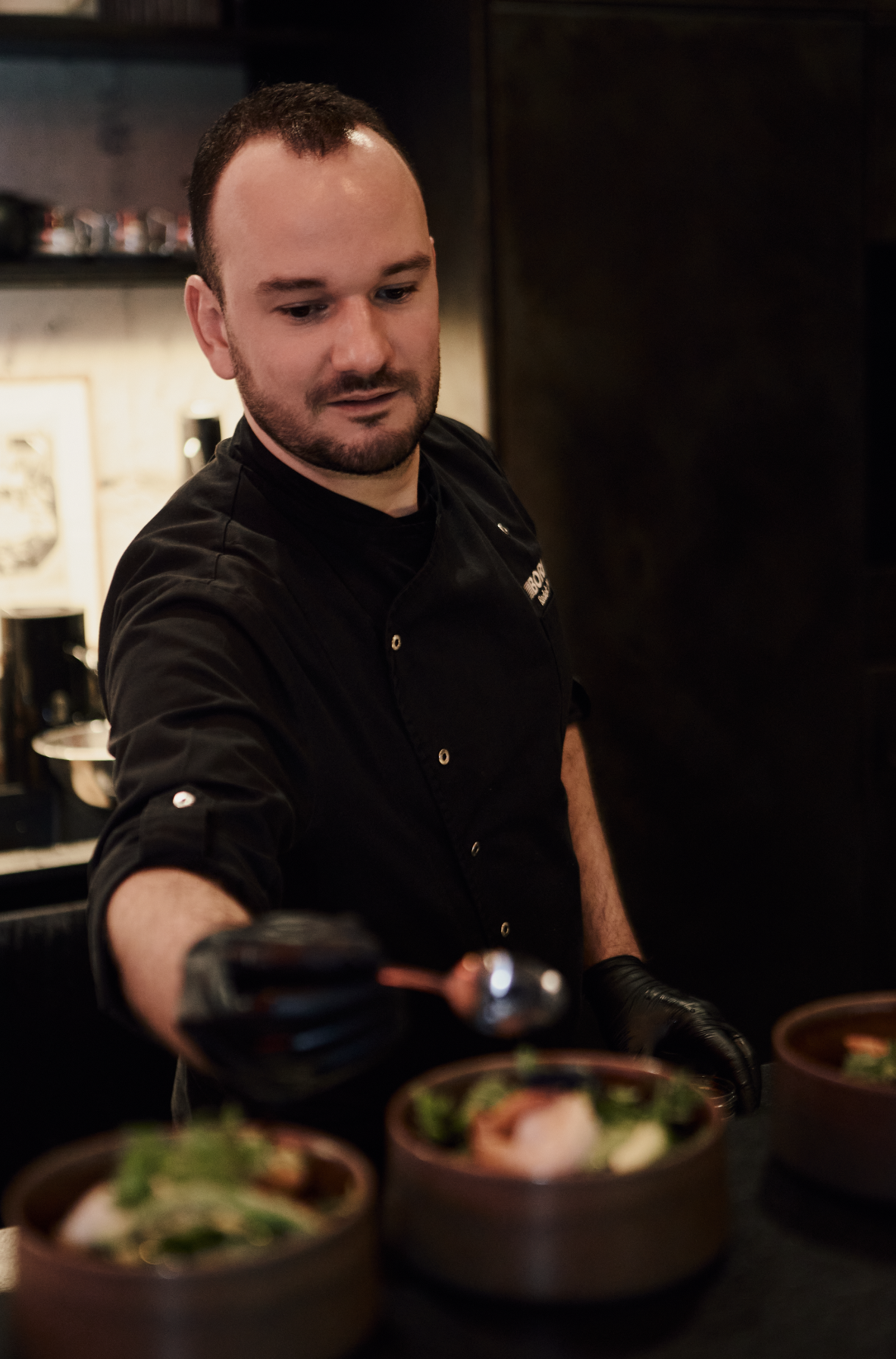 A male chef in a black uniform serving food into brown bowls at a restaurant.