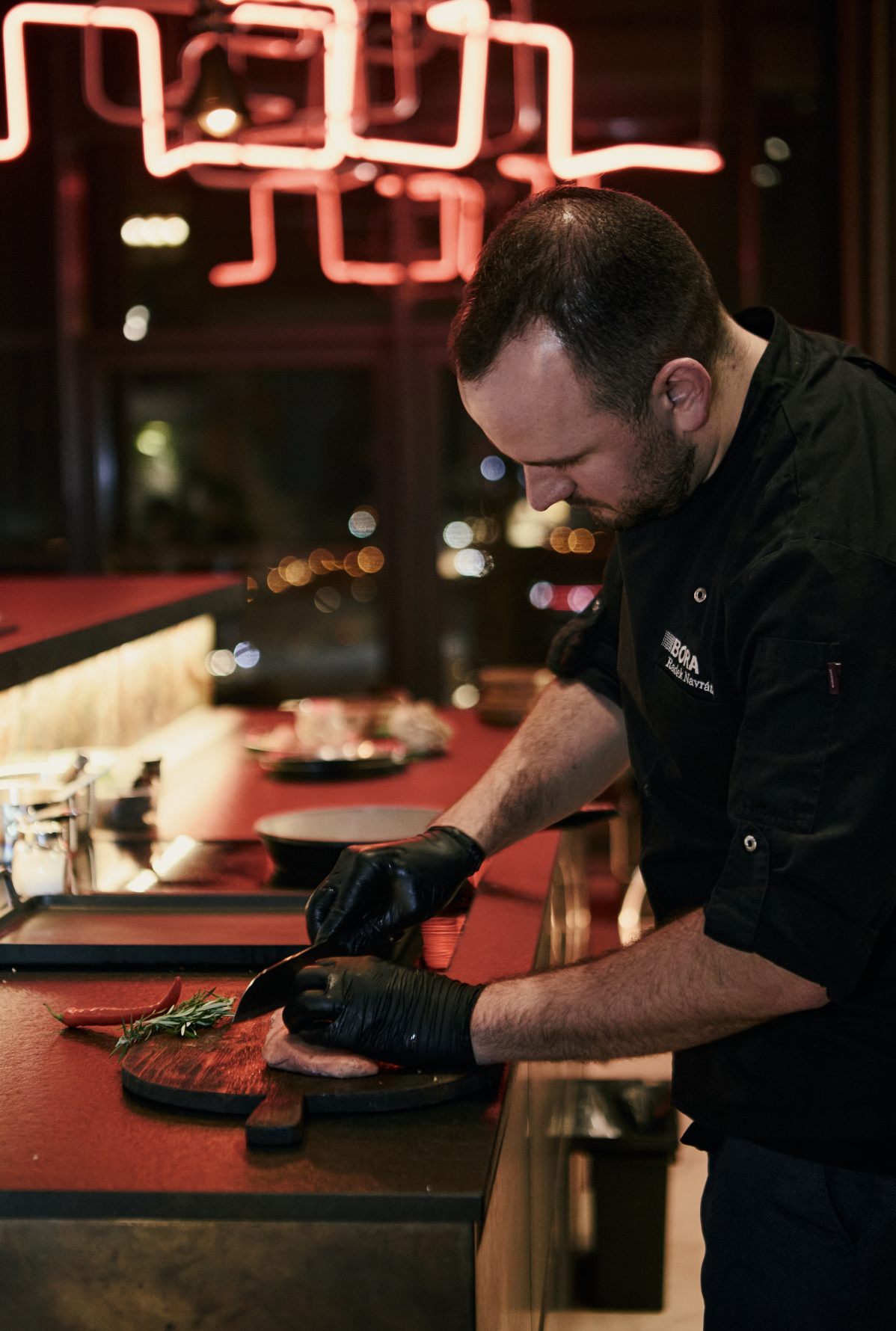 A chef in black uniform and black gloves preparing meat on a wooden cutting board in a kitchen with red countertops and a neon red sign in the background.