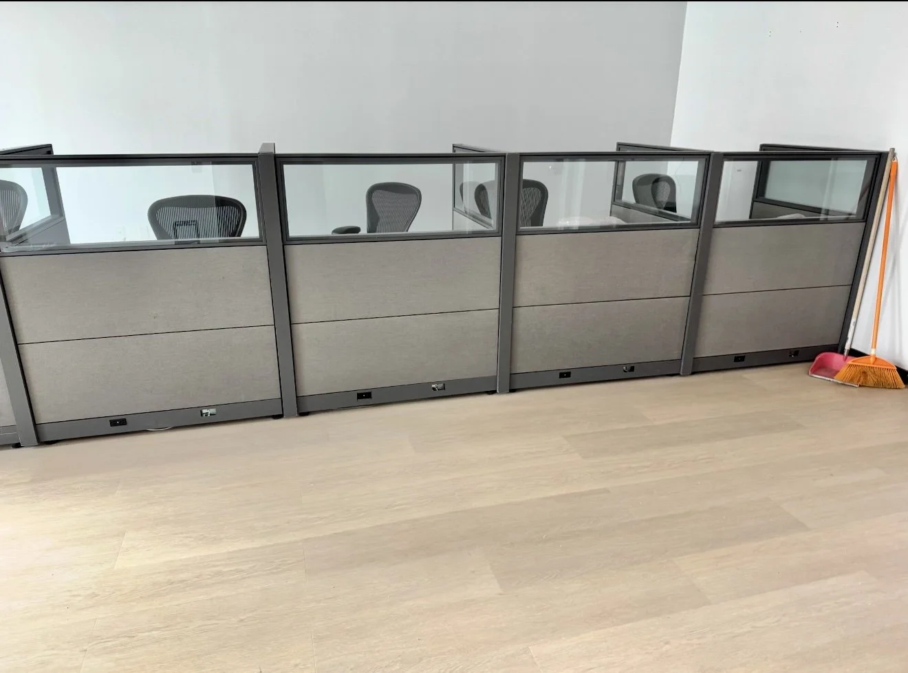 A row of office cubicles with glass tops and fabric panels, positioned against a plain wall, with a broom and dustpan in the corner on a light wood floor.