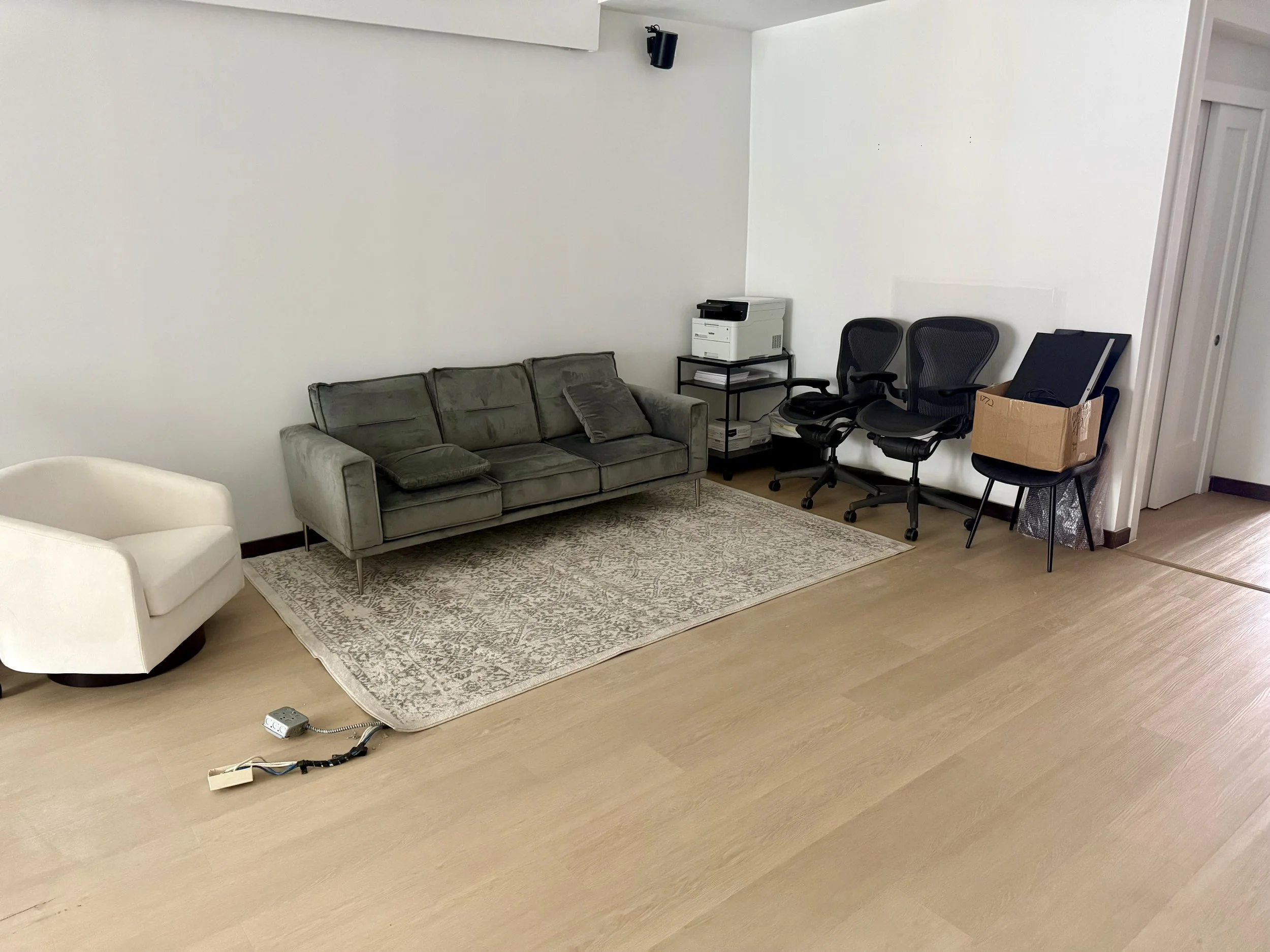 Office space with a gray couch, white armchair, and two black office chairs. There's a small table with a printer on top, a box of electronics, and some papers. The floor is light wood, and there is a power strip with cords on the floor. The wall is 