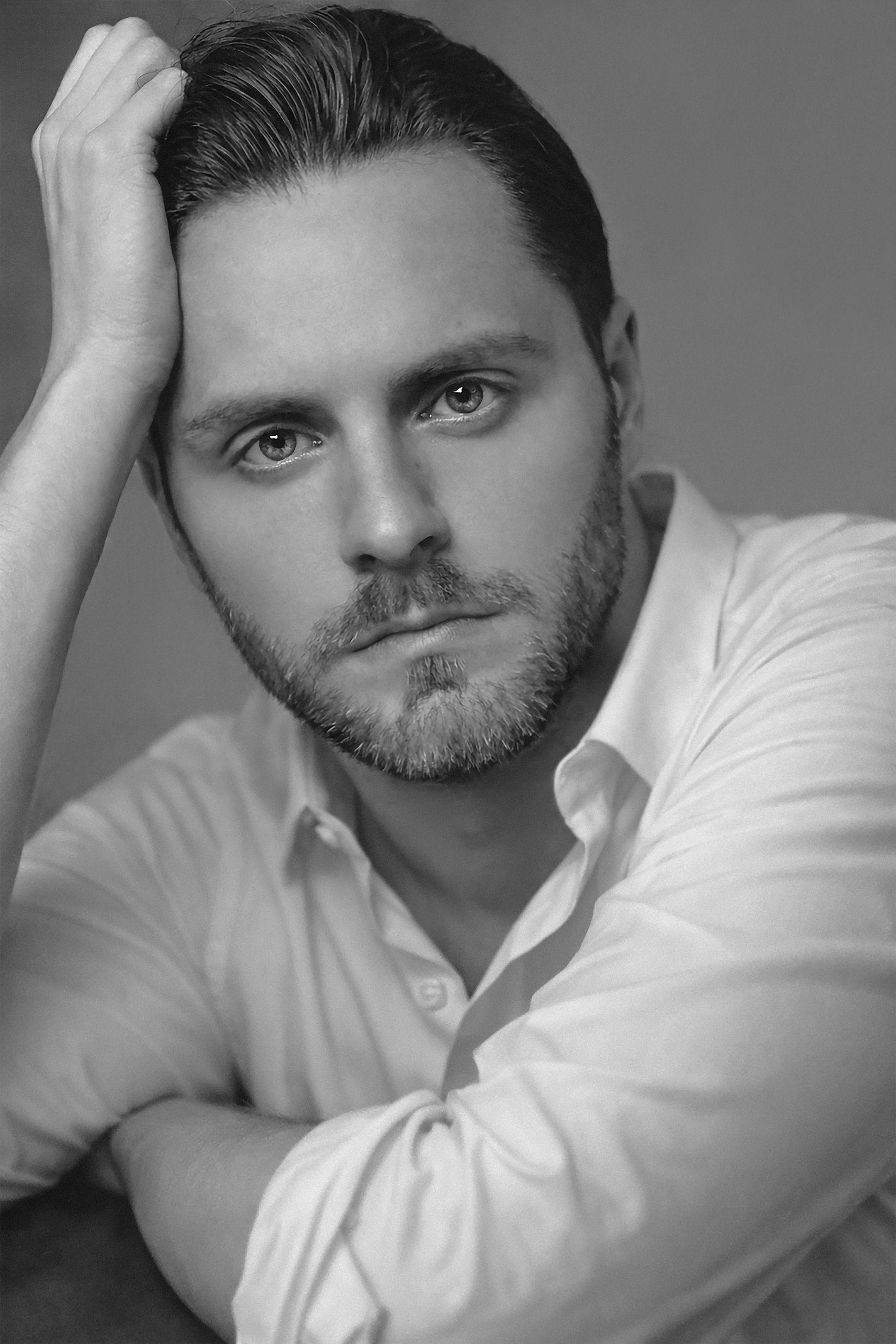 Black and white portrait of a man with short, styled hair and a beard, wearing a light-colored shirt and resting his head on his hand.