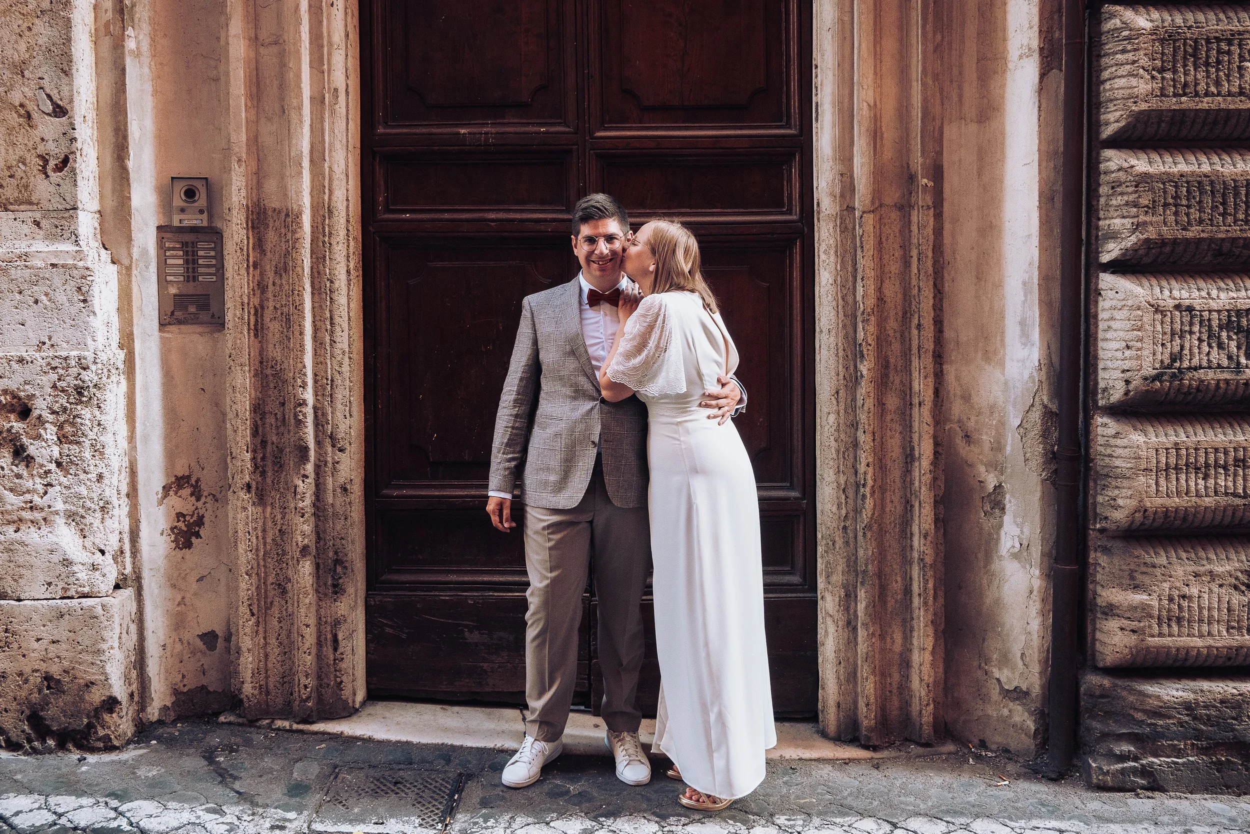 A man in a gray plaid suit and a woman in a white dress share a kiss in front of a large dark wooden door on a historic stone building.