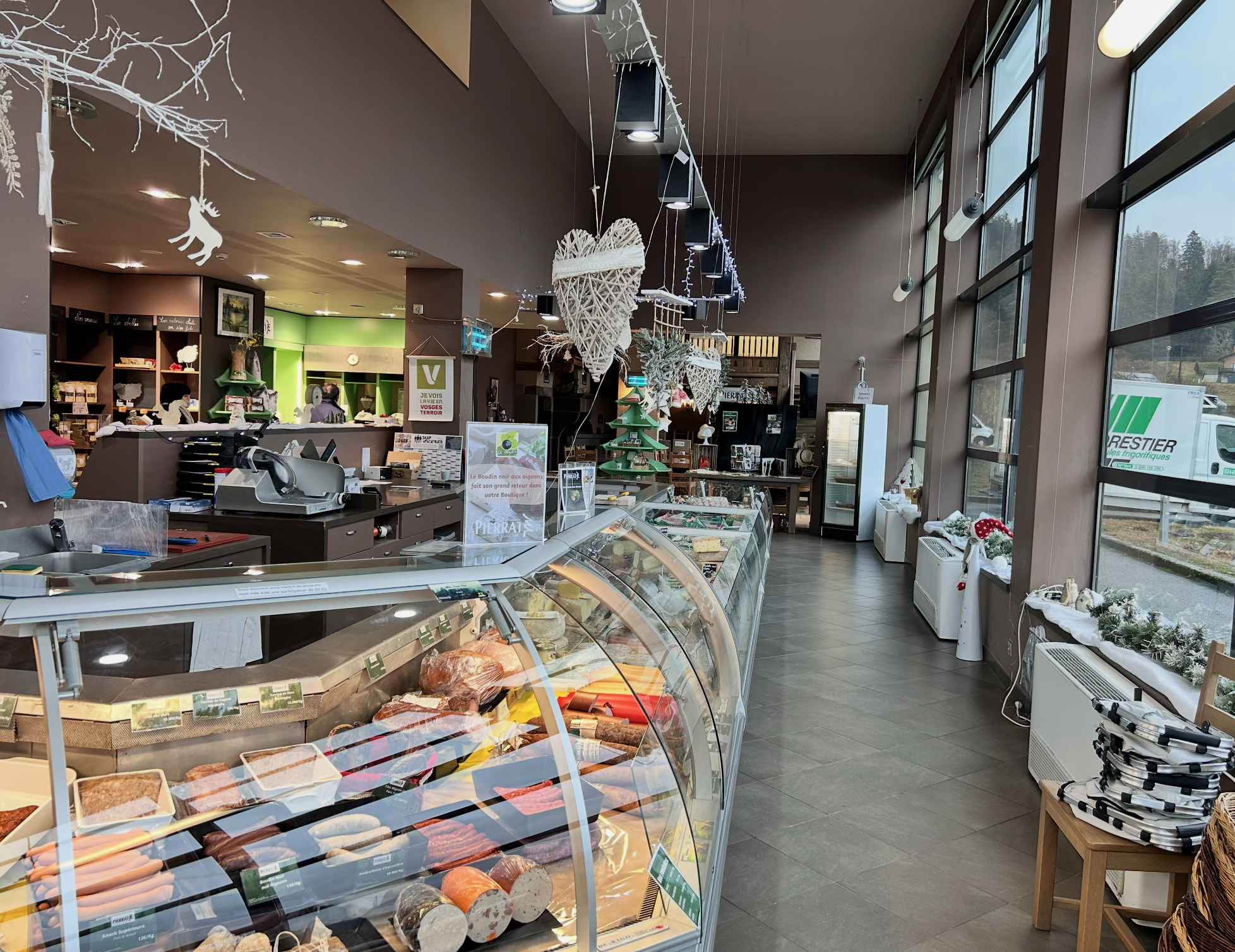 Intérieur d'une boucherie ou charcuterie avec un comptoir de viandes en premier plan, décorations de Noël suspendues au plafond, grande fenêtre laissant entrer la lumière naturelle côté droit, personnel en arrière-plan, étagères vertes et décorations festives.