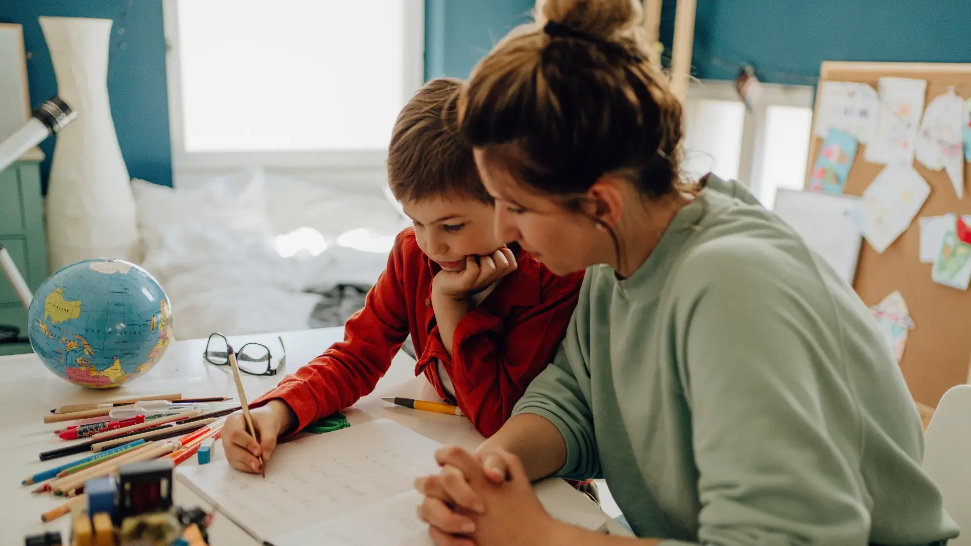 En voksen og barn sidder sammen ved et bord, de arbejder på en opgave eller tegning, omgivet af farveblyanter og skolematerialer, med en globus og briller på bordet.