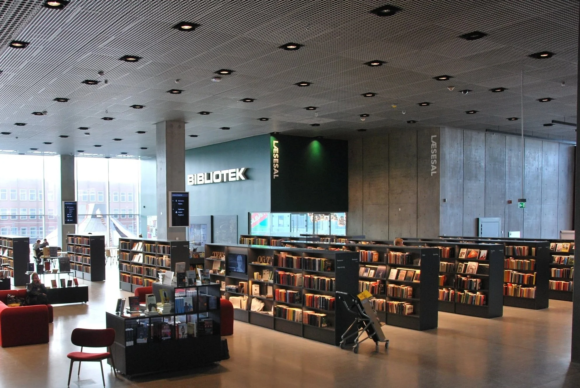 Indendørs billede af et bibliotek med bøger på hylder, læsekroge med røde stole og store vinduer, hvor der er naturligt lys. Der er skilte med teksten 'BIBLIOTEK' og 'LÆSESAL'.