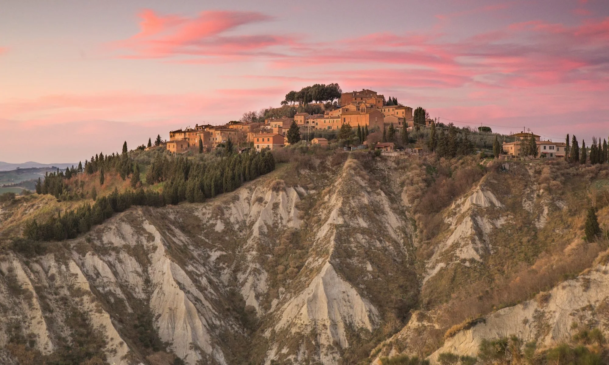 Un village perché sur une colline rocheuse avec des maisons en pierre, entouré d'arbres et sous un ciel rose au coucher du soleil.