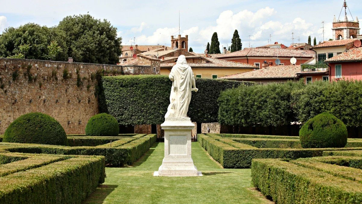 Une statue blanche d'une figure religieuse dans un jardin bien entretenu avec des buissons taillés et des arbres, avec des bâtiments résidentiels en arrière-plan.