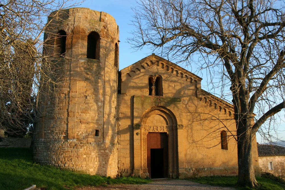 Une ancienne église en pierre avec une tour ronde et un toit en pente, entourée d'arbres sans feuilles, lors d’un coucher de soleil.