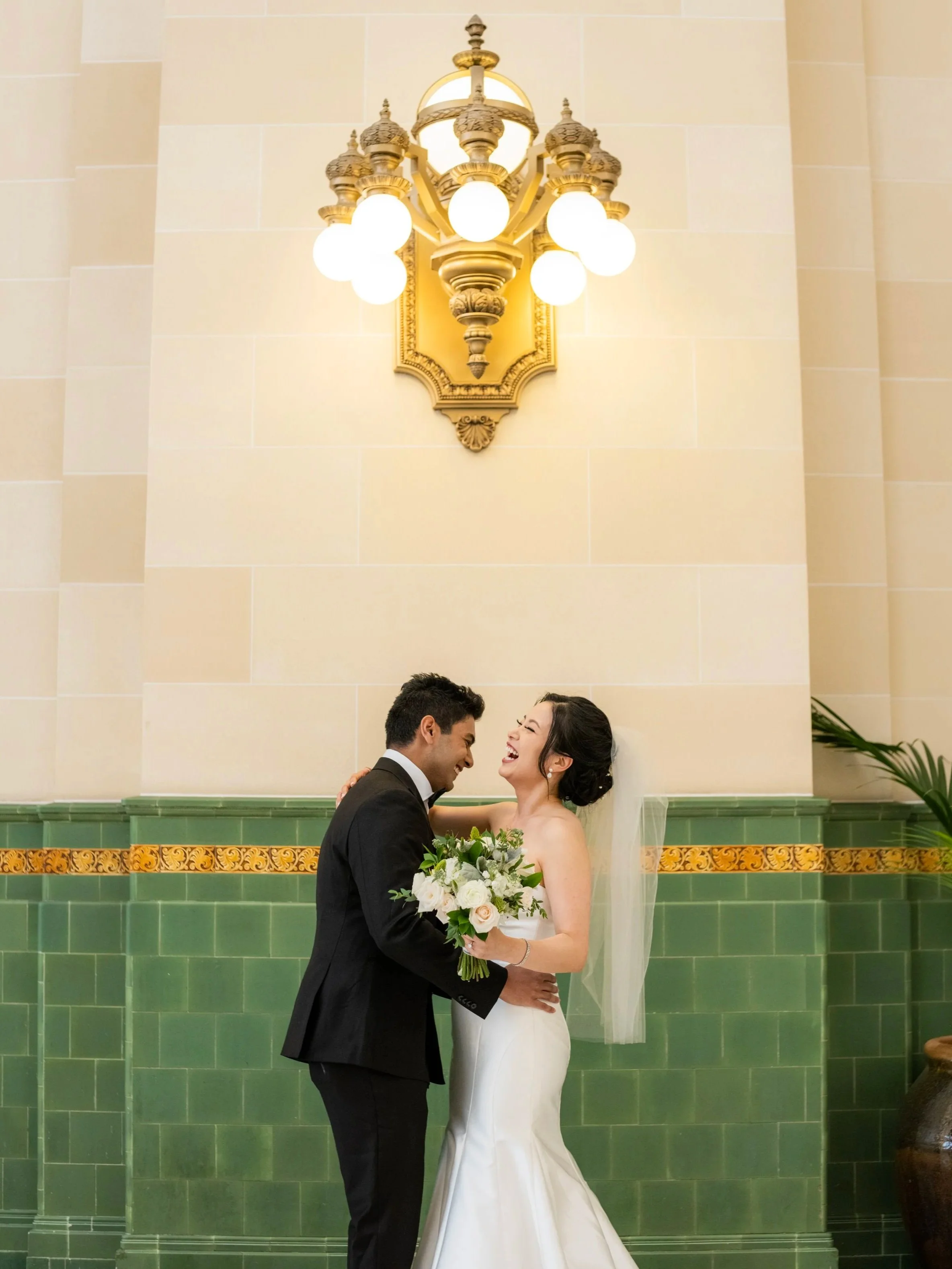 A bride and groom share a joyful moment during their wedding ceremony, standing close together in front of a green tiled wall with decorative yellow border, illuminated by an ornate chandelier overhead.