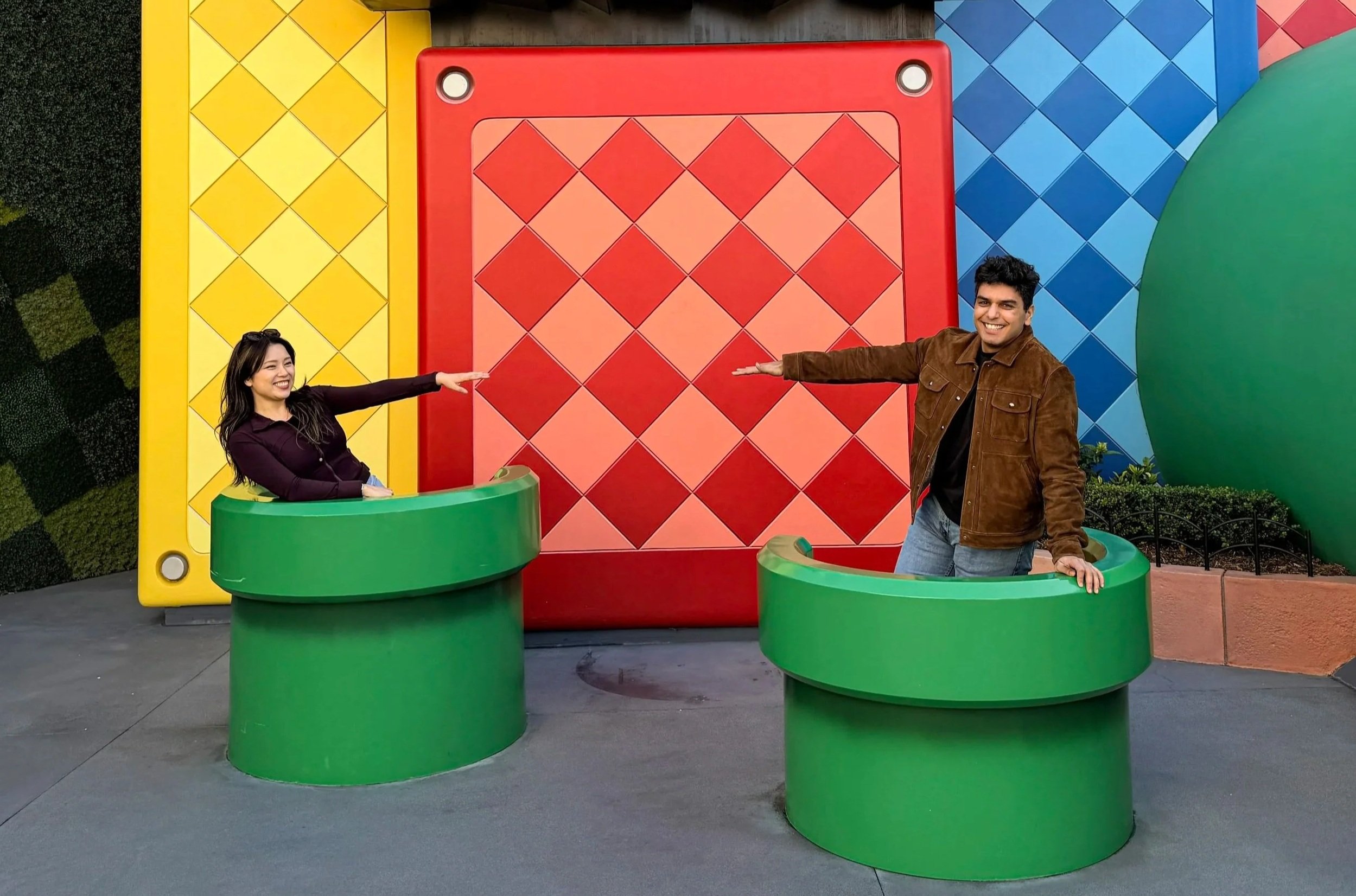 Two people, a woman and a man, standing inside large green circular structures in front of a colorful, patterned backdrop at an amusement park or entertainment venue. They are smiling and extending their arms towards each other.