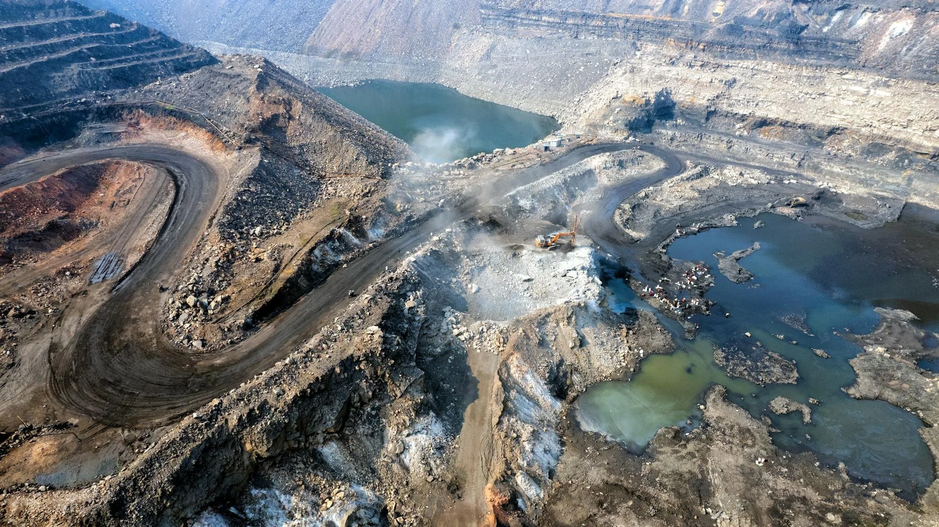 Aerial view of a mining operation with winding dirt roads, large excavations, water-filled pits, and a pond, amidst rocky terrain.