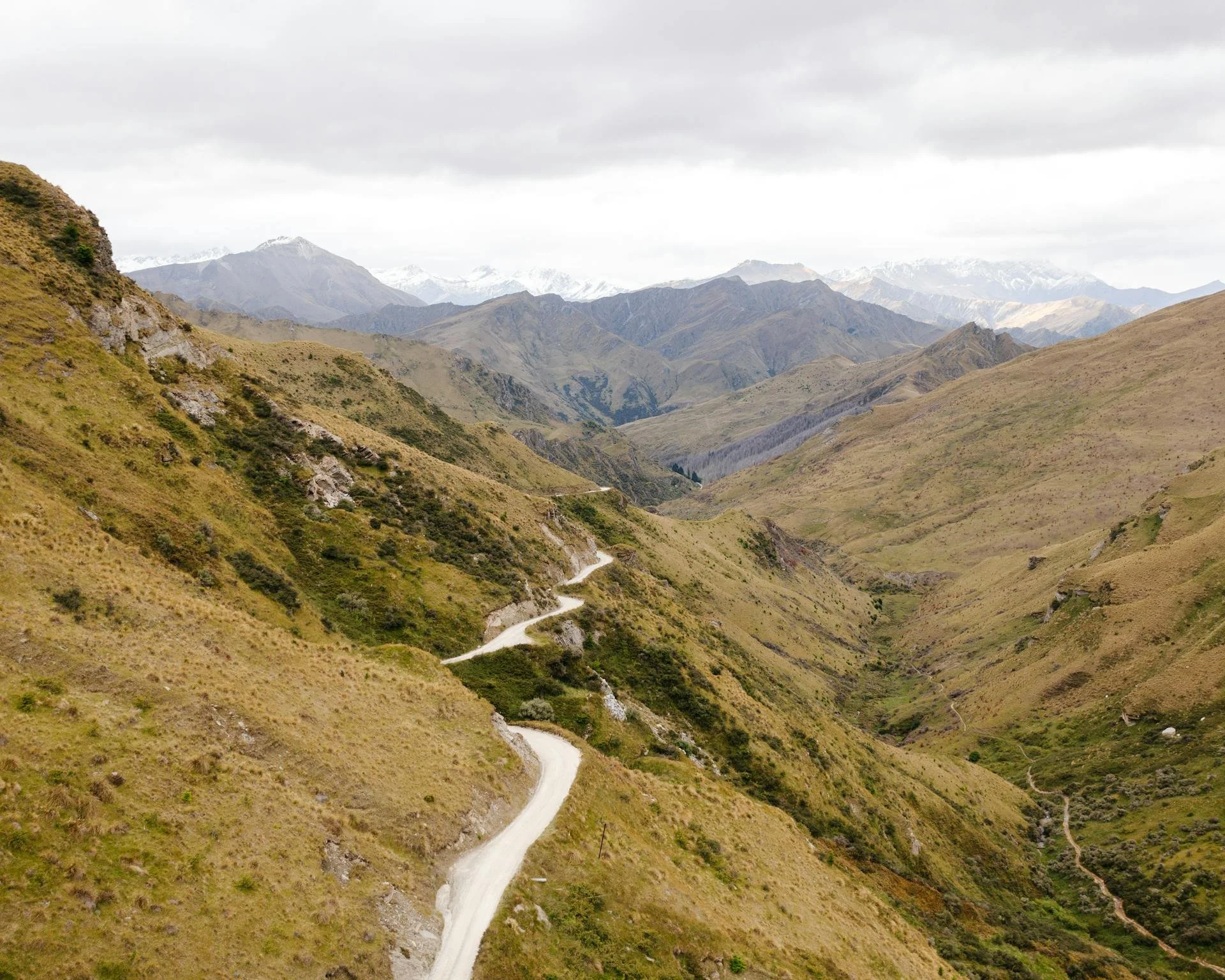 A winding dirt road runs through a green and brown mountainous landscape with distant snow-capped peaks under a cloudy sky.