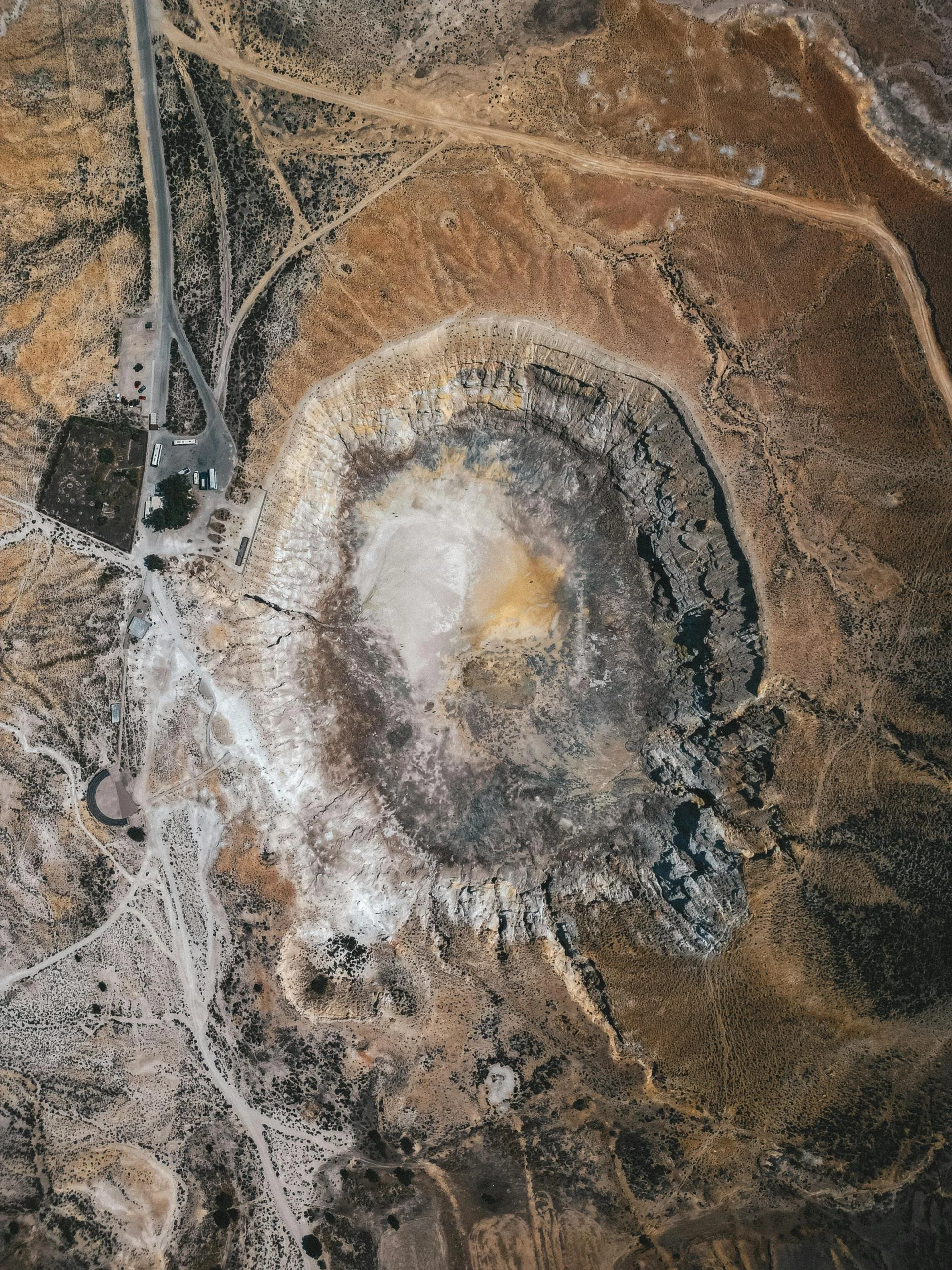 Aerial view of a large open-pit mining or quarrying operation, with roads and facilities on the left side and a large excavated crater in the center surrounded by rugged terrain.