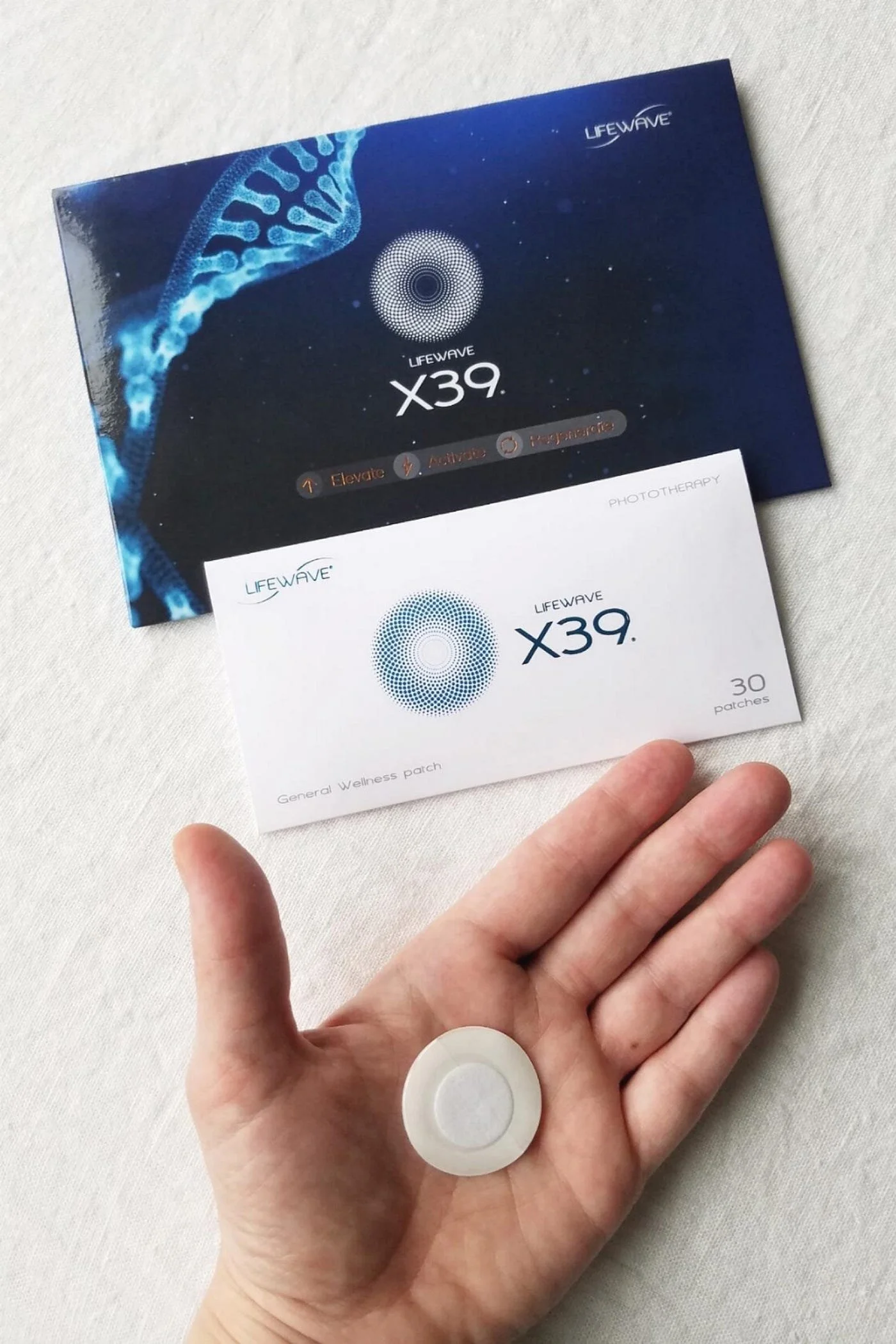 A hand holding a round, white patch with a gray circle in the center, next to packaging and a pamphlet for LIFEWAVE X39 patches.