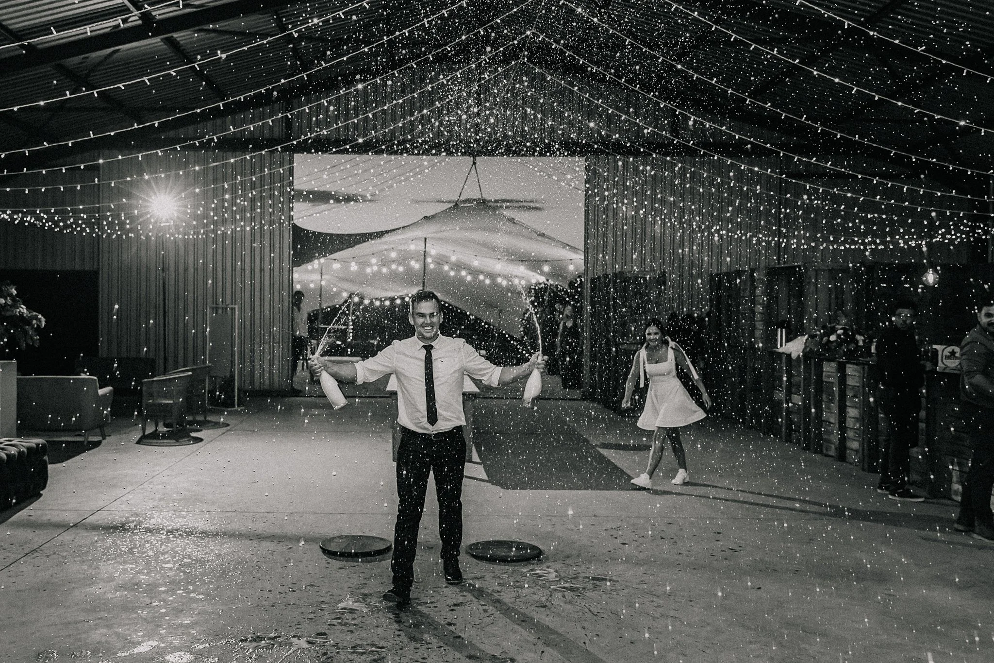 A man in a white shirt and loosened tie, smiling, celebrating with two champagne bottles, amid a shower of confetti or snow inside a decorated venue with string lights and group of people in the background.