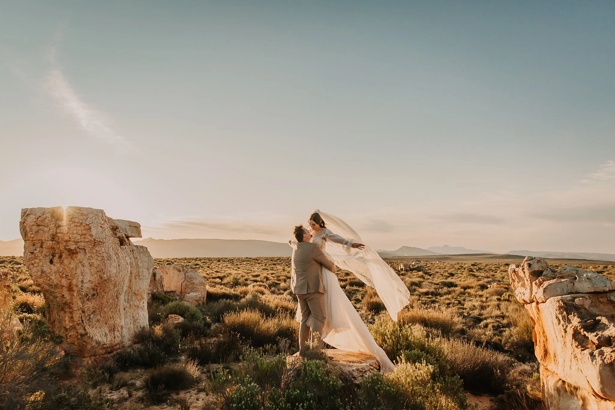 Groom is lifting bride while she is holding on to her veil which is blowing in the wind.