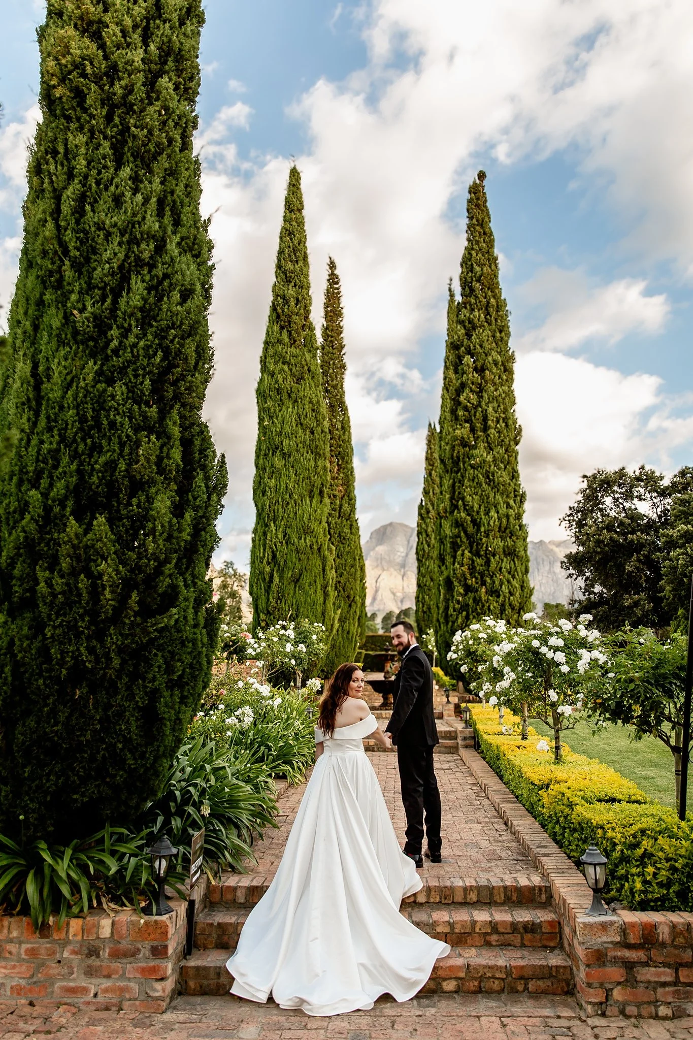 A bride and groom holding hands on a brick pathway surrounded by greenery and white flowers, with tall cypress trees and mountains in the background during daytime.