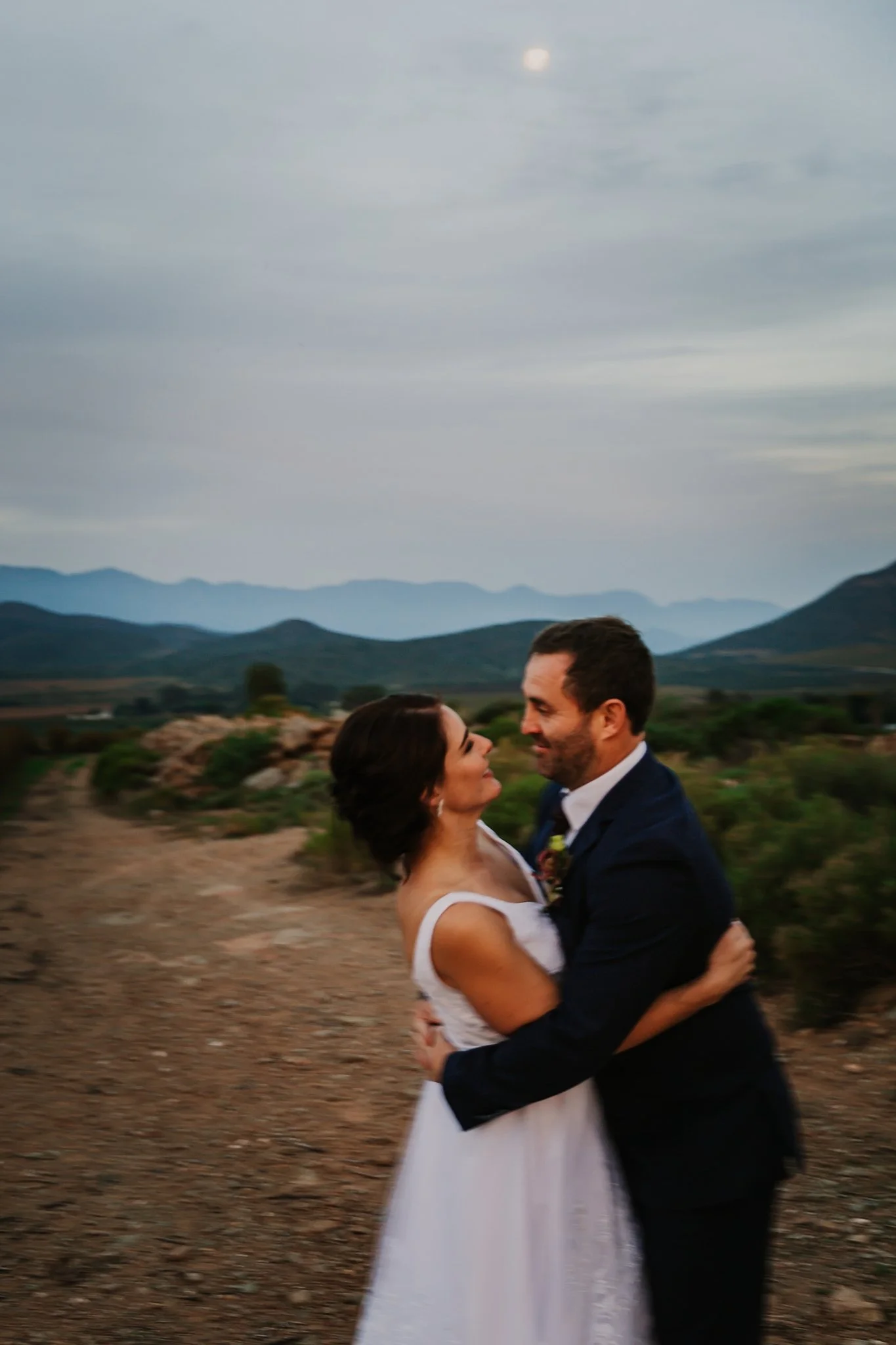 A man and woman in wedding attire embracing outdoors on a dirt path with mountains in the background at dusk.
