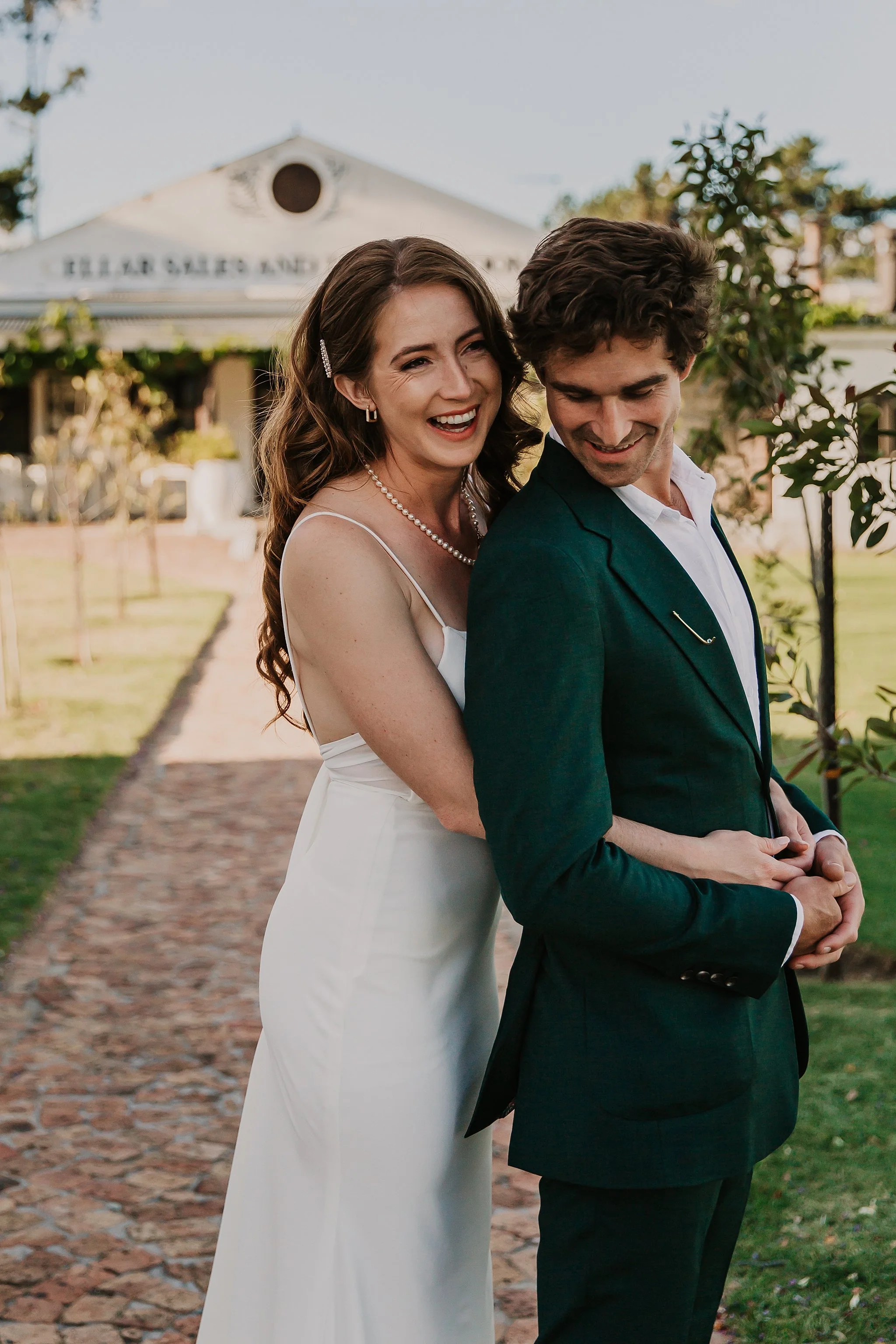 A smiling woman in a white dress embraces a man in a dark green suit outside a building with a sign that says 'Heaven Salads and...' in the background.
