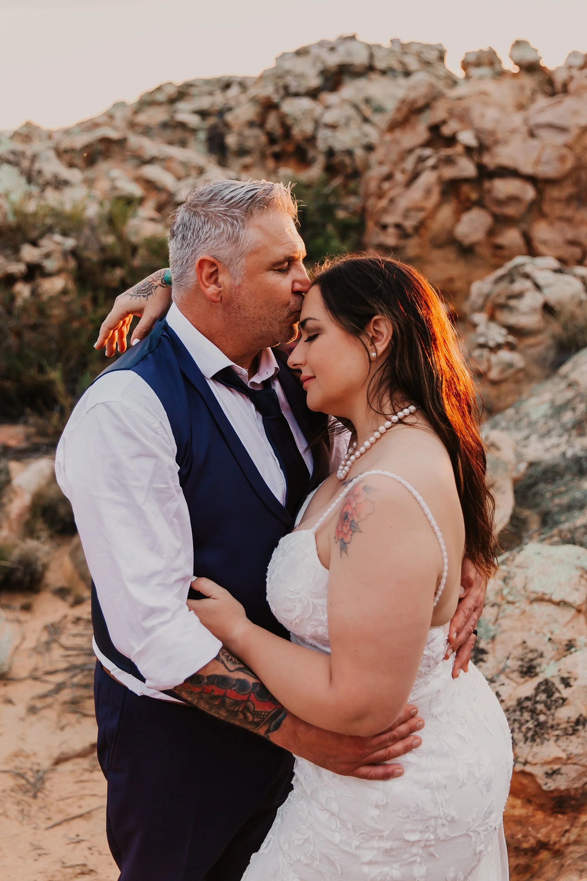 A couple dressed in wedding attire embracing outdoors during sunset, with rocky terrain in the background.