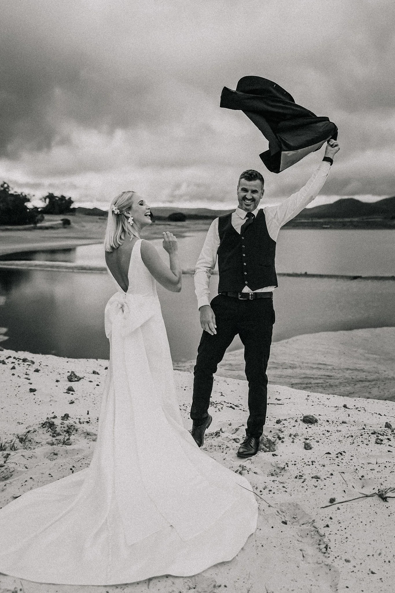 A bride and groom celebrating outdoors near a body of water, with the groom holding a suit jacket in the air and both smiling.
