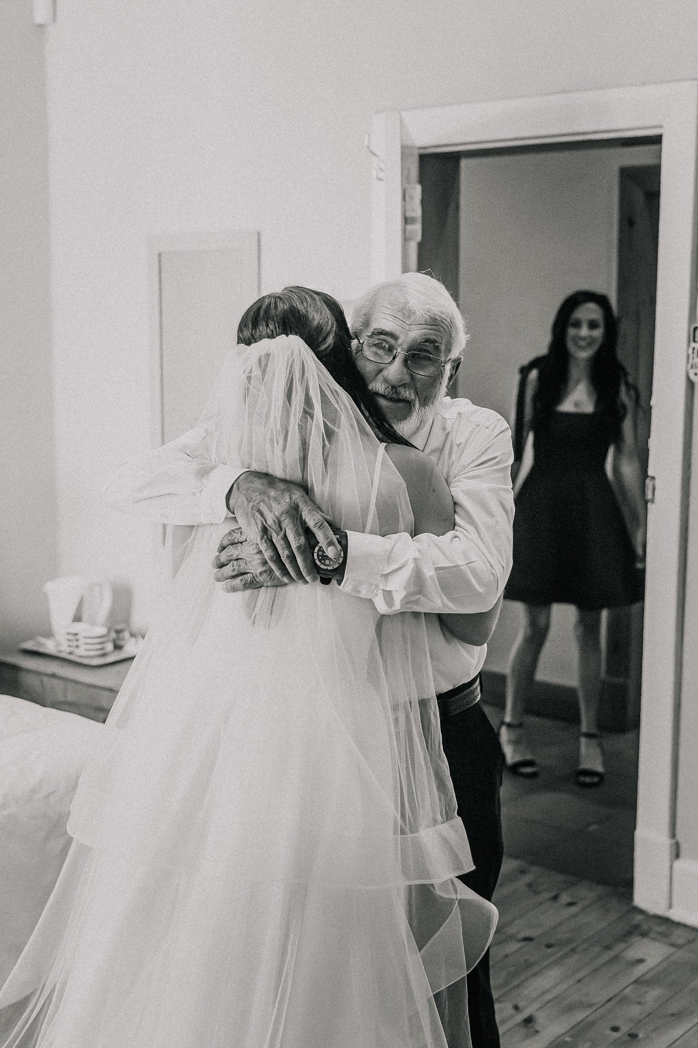 An elderly man in a white shirt embraces a woman in a wedding dress, who is wearing a veil, in a room. A younger woman in a black dress and heels stands in the background, smiling and watching the scene.