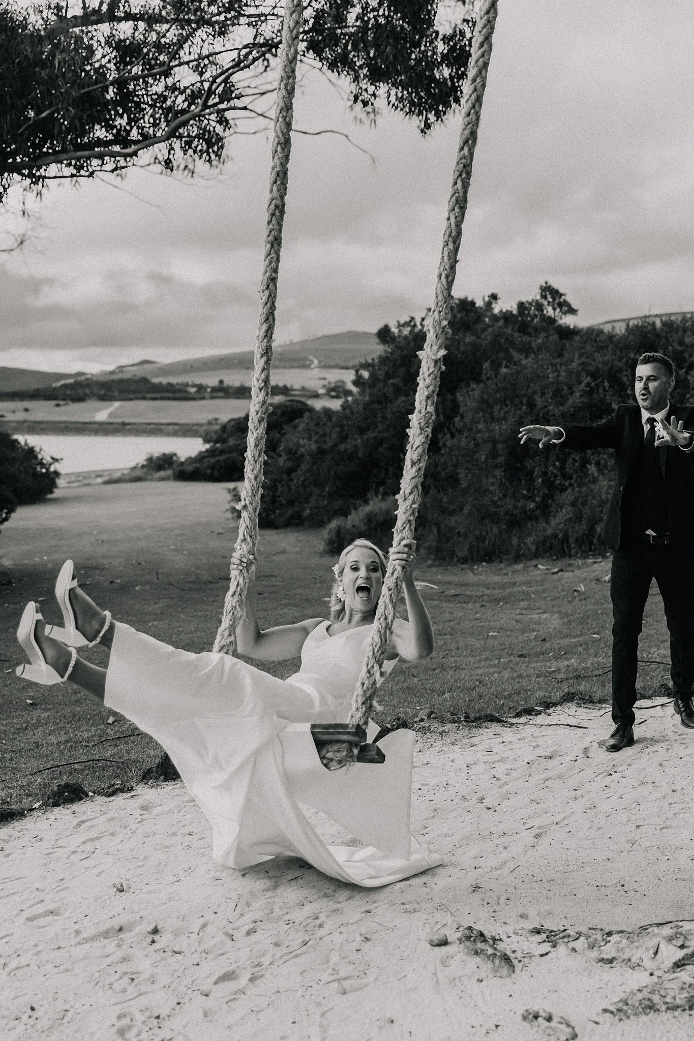 A woman in a wedding dress on a swing, smiling and holding the ropes, with a man in a suit standing nearby, outdoors in a scenic landscape.