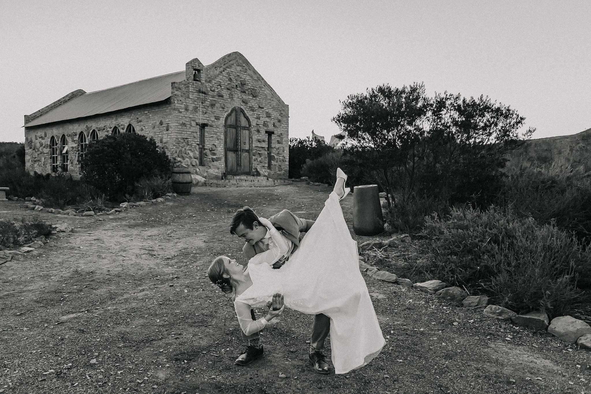 A black-and-white photo of a man in a suit and a woman in a wedding dress dancing outside near a stone church building, surrounded by bushes and trees.