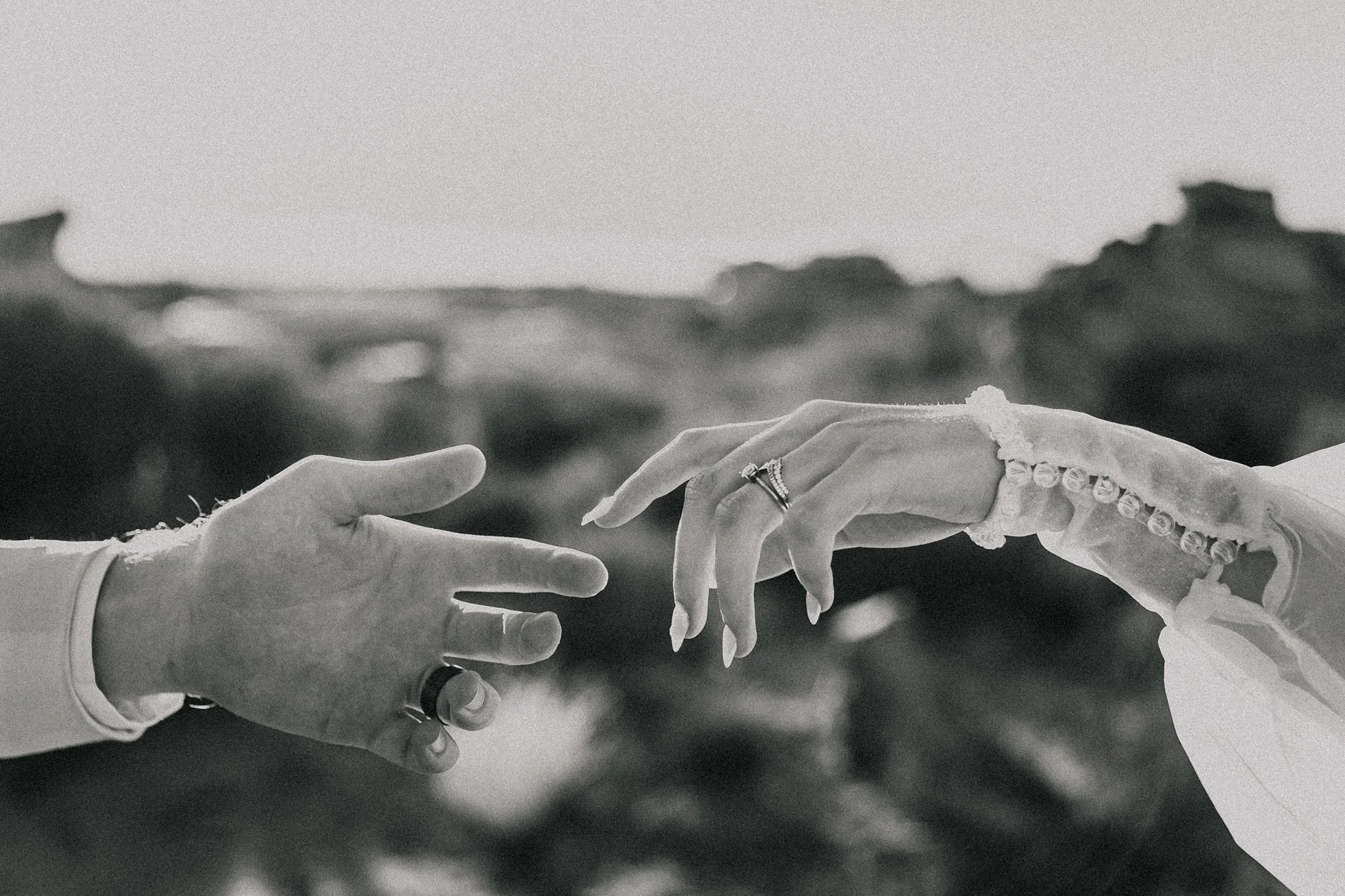 Black and white photo of two hands reaching towards each other, with one hand on the left and the other on the right, both wearing rings. The background is blurred, suggesting an outdoor setting.