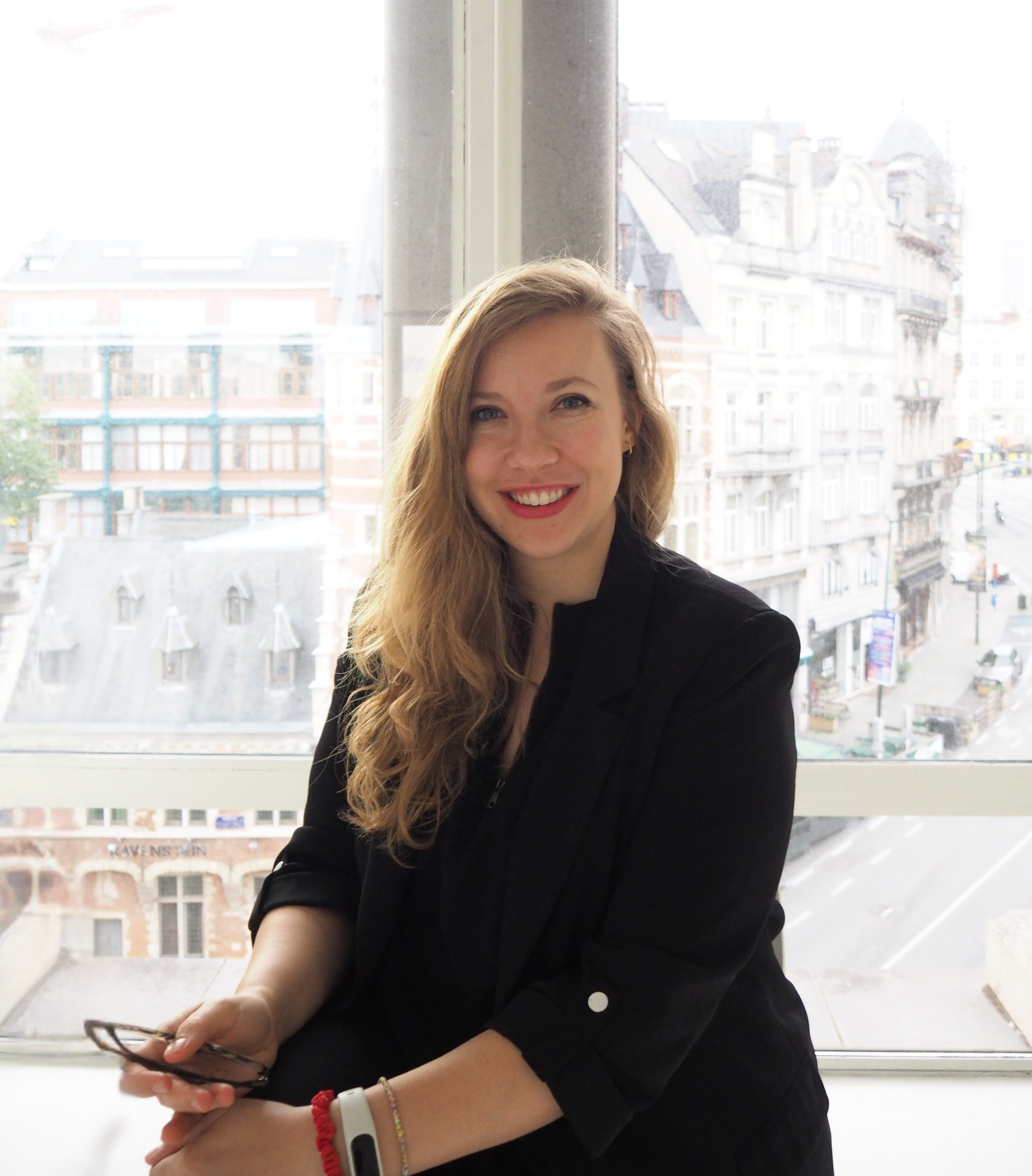 A woman with long wavy blonde hair smiling, wearing a black blazer, sitting in front of a large window overlooking city buildings.