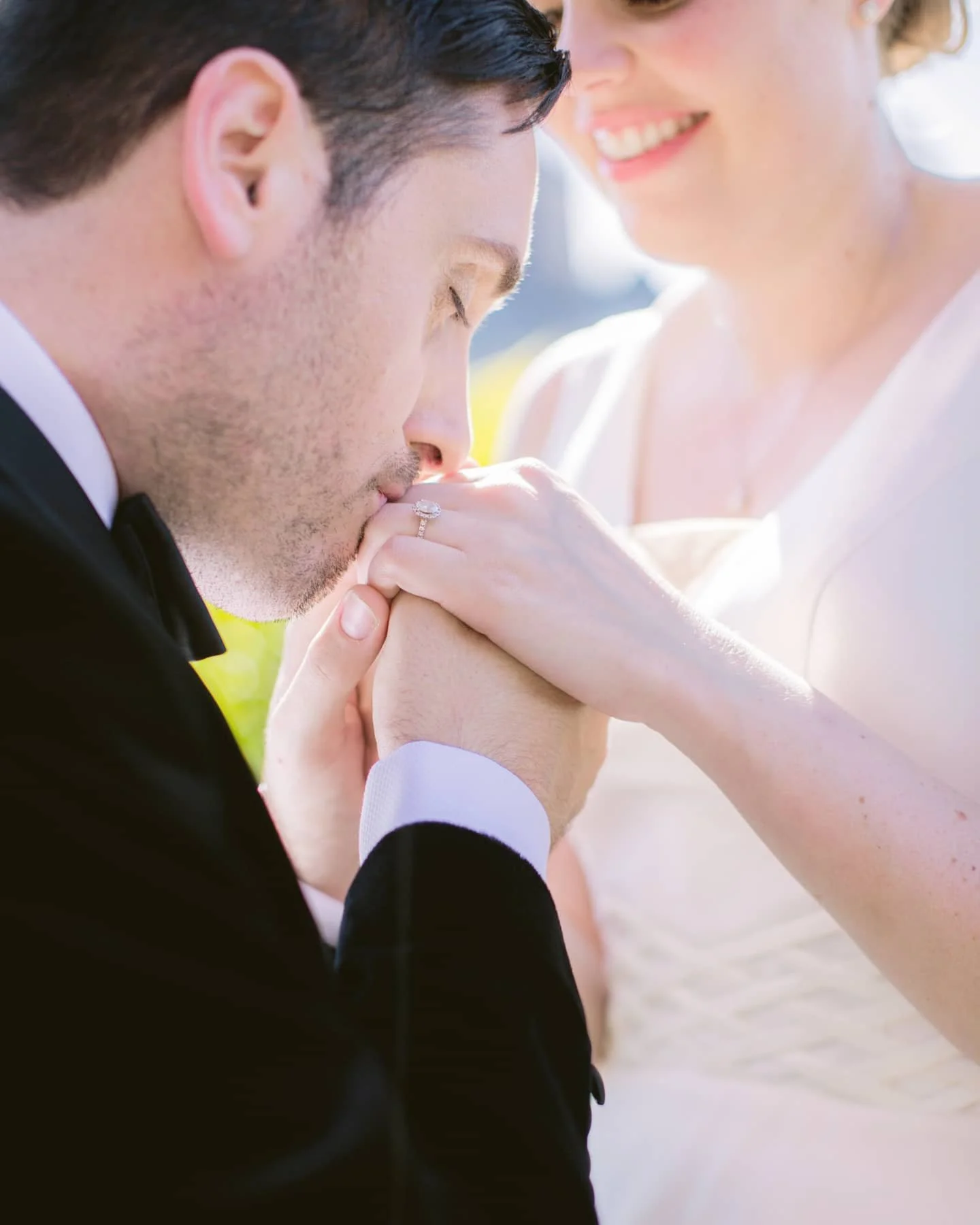Un homme en costume noir et une femme en robe élégante lors d'un mariage, où l'homme embrasse la main de la femme, qui porte une bague de fiançailles.
