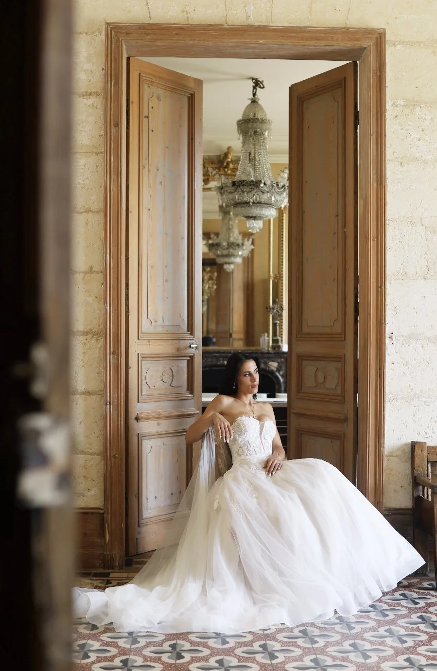 Jeune femme en robe de mariée assise dans une pièce élégante au château Fengari avec lustres en cristal et parquet en carreaux décoratifs. 