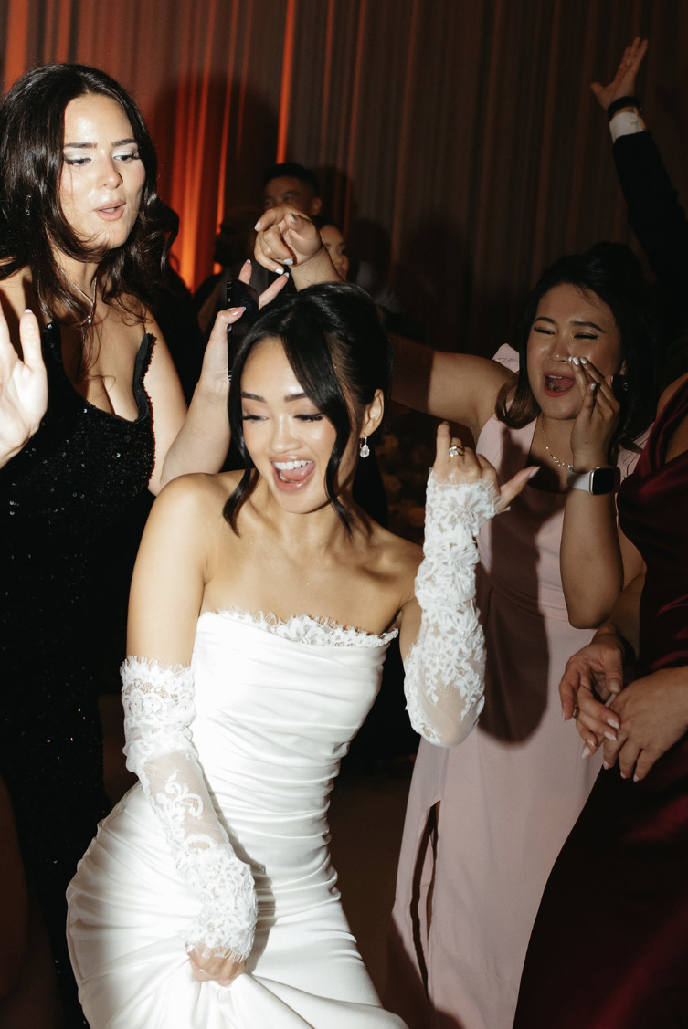 Des femmes dansant et souriant à une fête ou un mariage, avec une femme en robe de mariée blanche au centre. Photo de soirée de mariage style papparazzi.