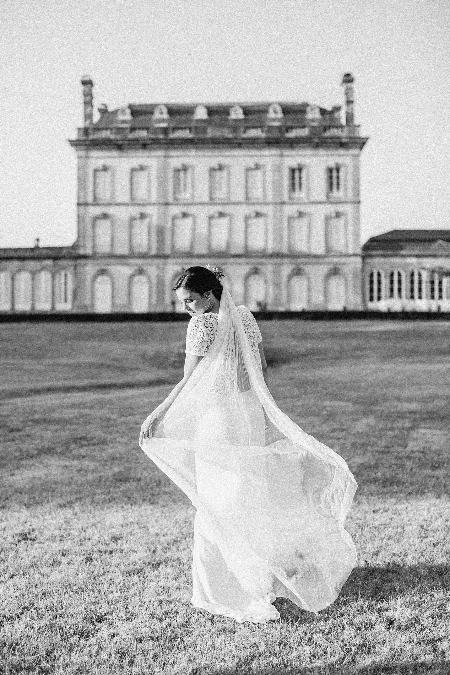 Une mariée en robe de mariage, tenant son voile, sur une pelouse devant un grand château en France.
