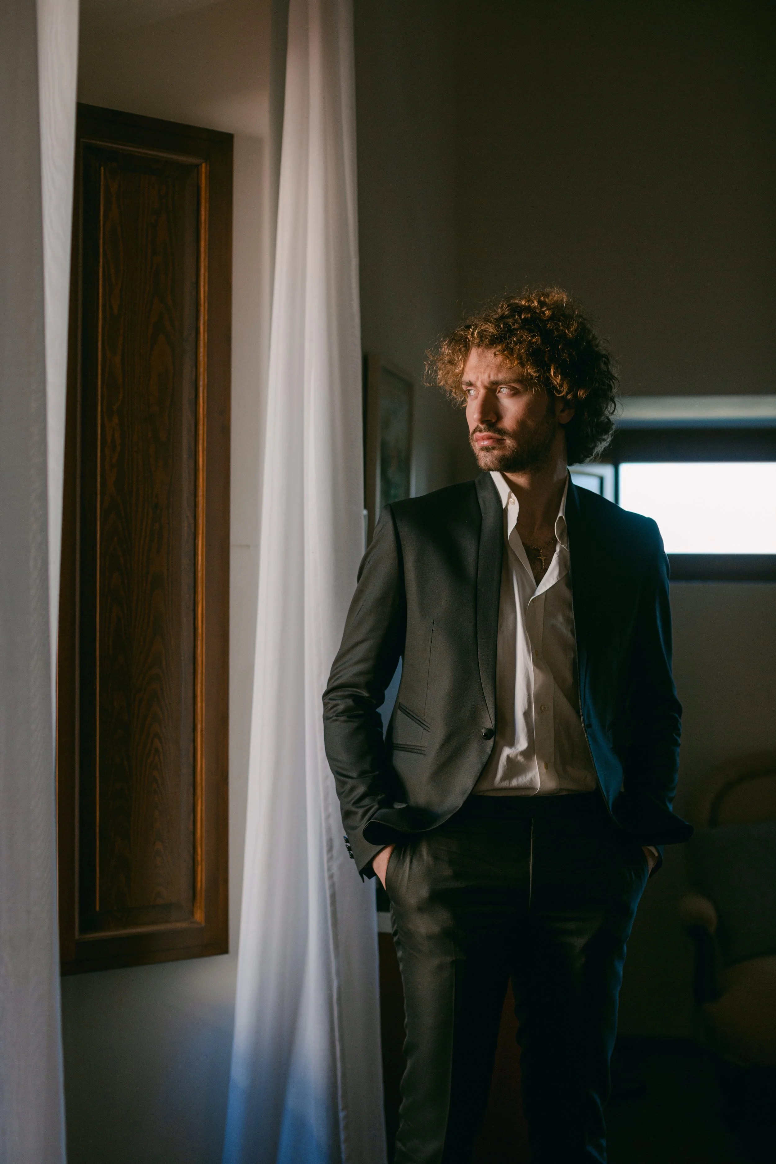 Futur marié en Italie en costume noir et chemise blanche se tient dans une pièce, regardant par une fenêtre, avec des mains dans les poches.