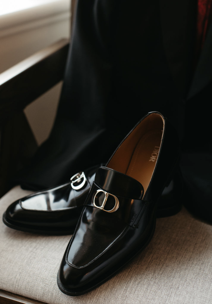 Chaussures de luxe noires en cuir verni avec boucle dorée 'Dior' sur une surface beige, en arrière-plan un fauteuil en bois.