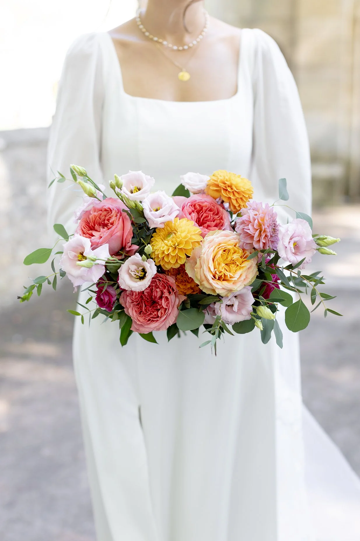 Femme en robe blanche tenante un bouquet de fleurs colorées, comprenant des roses, des dahlias et des lis, avec un collier de perles et un pendentif doré.