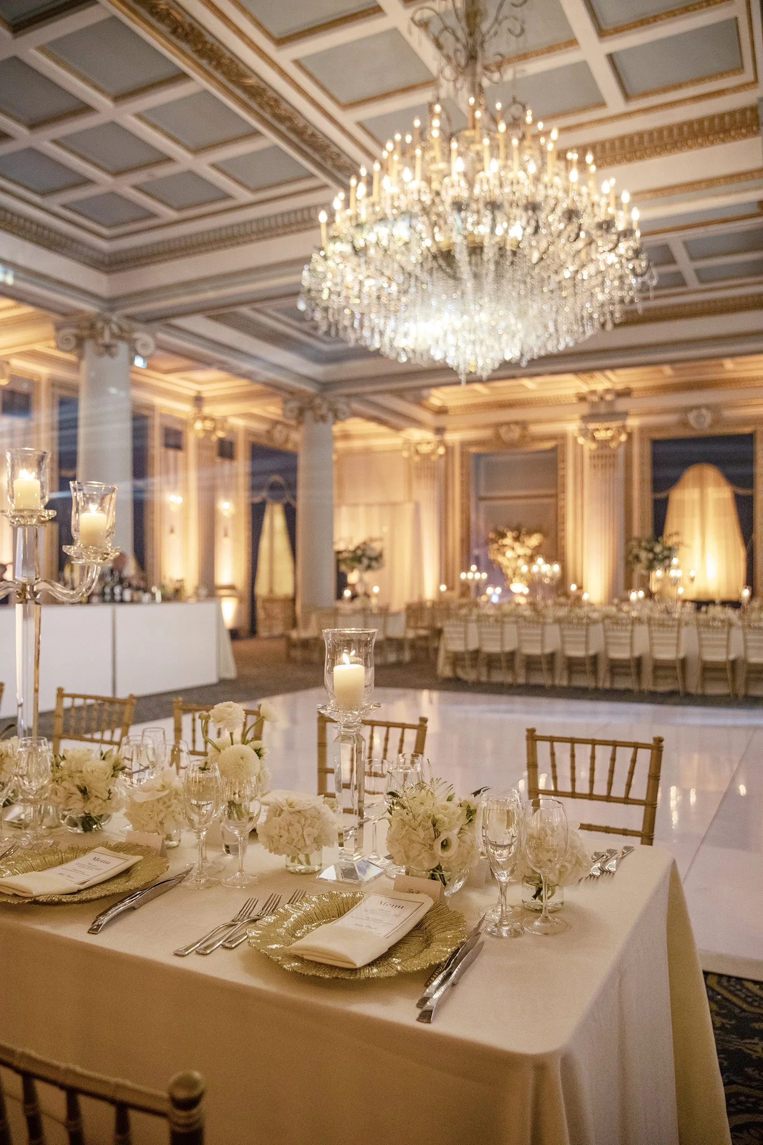 Salle de réception élégante au Ritz avec plafond orné de moulures, grand chandelier en cristal, table de mariage décorée de fleurs blanches, bougies et vaisselle raffinée, ambiance chaleureuse et sophistiquée.