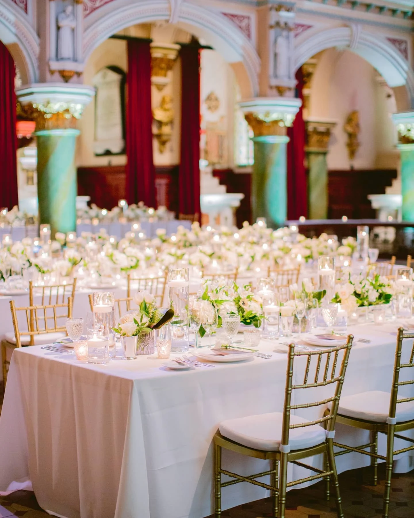 Salle de réception élégante avec une grande table dressée pour un événement, décorée de fleurs blanches et de bougies. Arches ornées, colonnes vertes et rideaux rouges en arrière-plan.