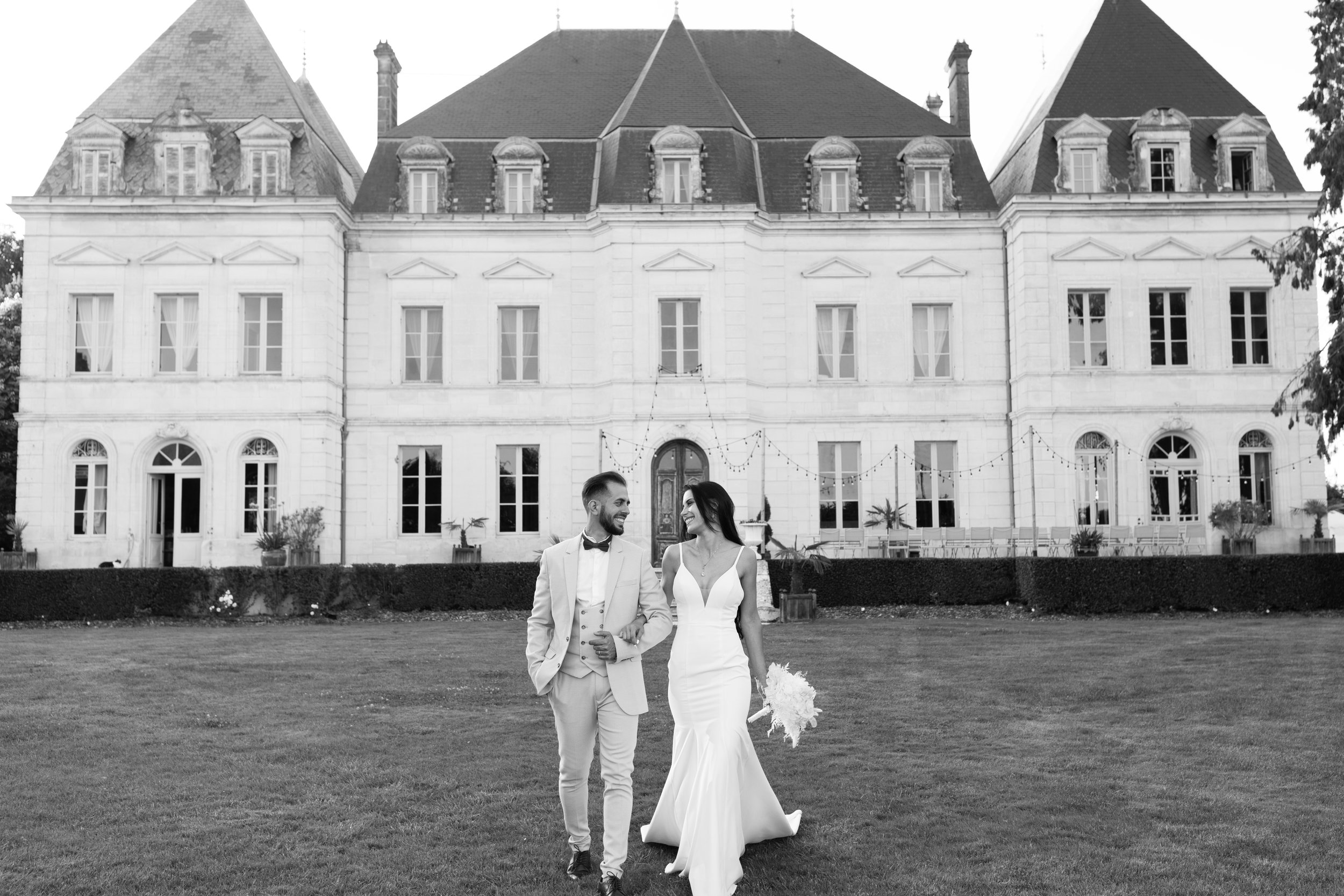 Un couple de mariés marche sur l'herbe devant le château Fengari en France lors de leur mariage, décoré avec des guirlandes lumineuses.