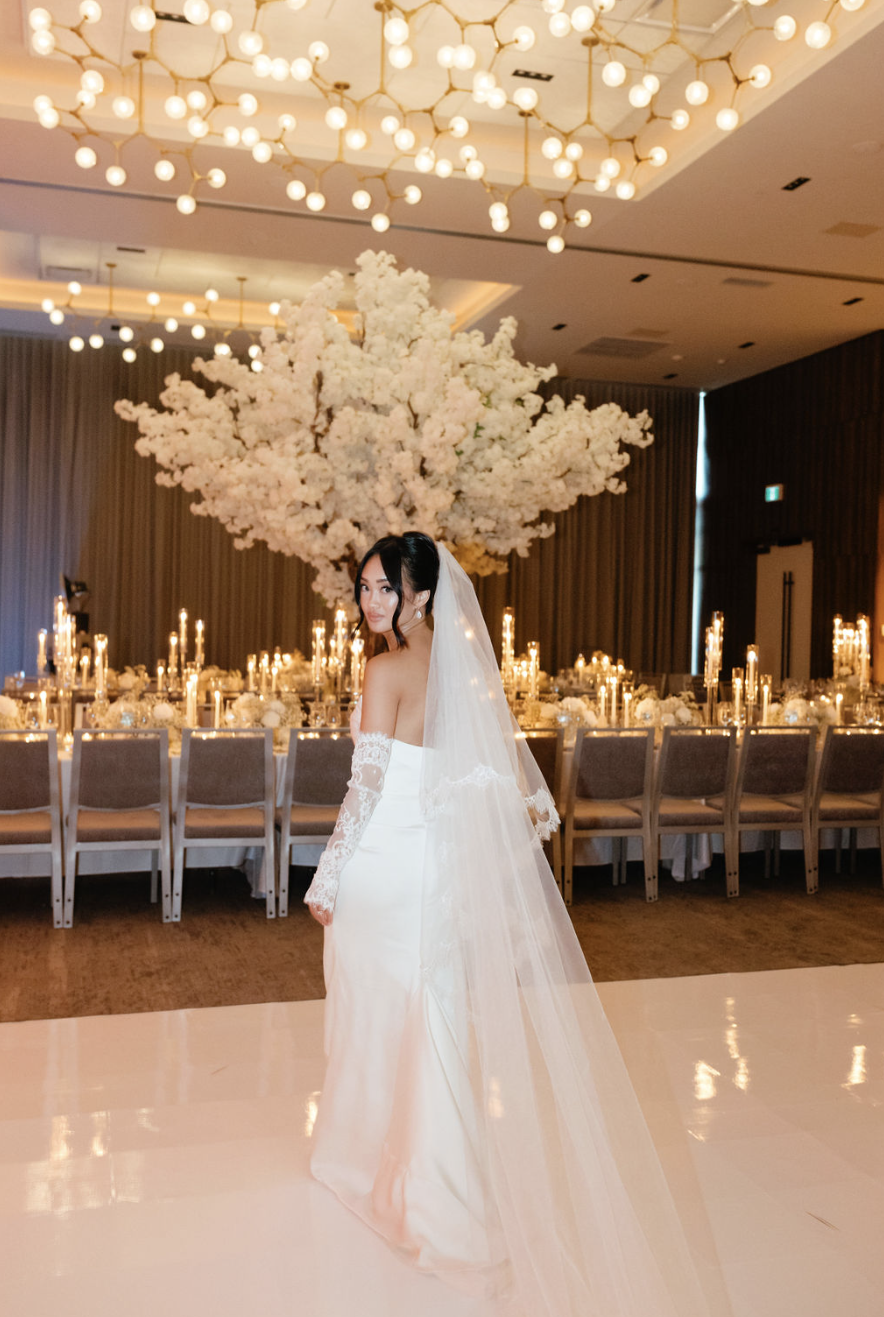 Une mariée en robe blanche avec un voile dans une salle élégante décorée de chandeliers et d'un grand arbre floral blanc au fond. Photo de soirée de mariage style papparazzi.