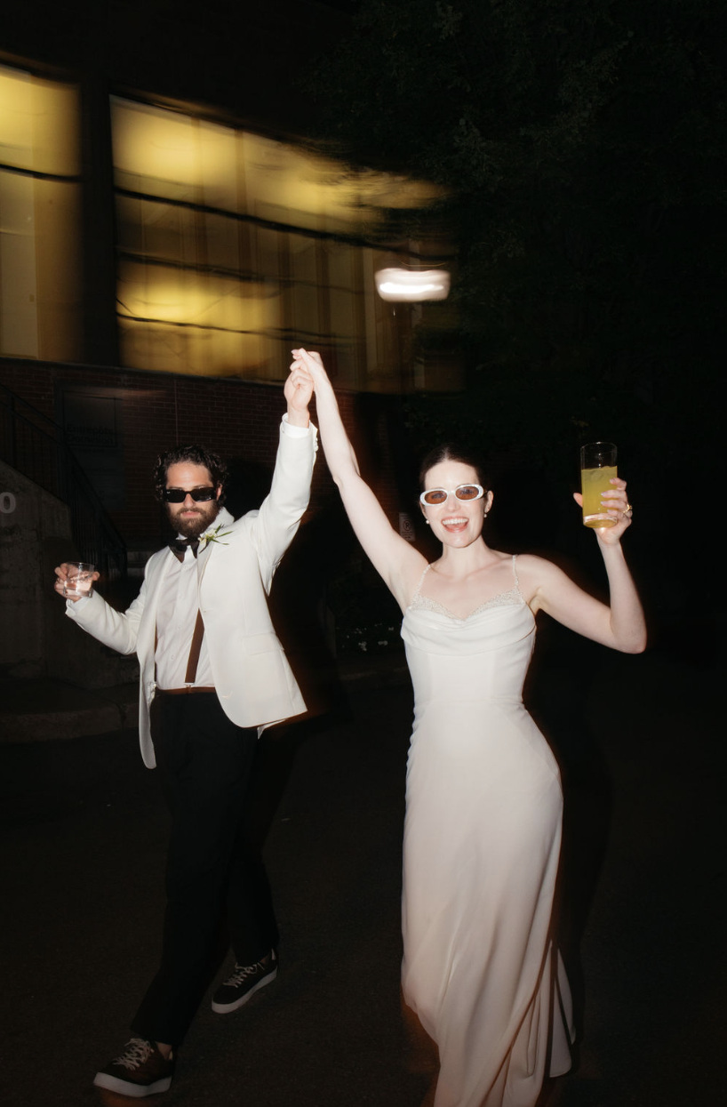 Un couple habillé élégamment, levant les mains en dansant ou célébrant, la femme porte une robe blanche longue et des lunettes de soleil, l'homme porte un costume blanc avec une chemise noire et des lunettes de soleil, lors d'une fête nocturne avec d